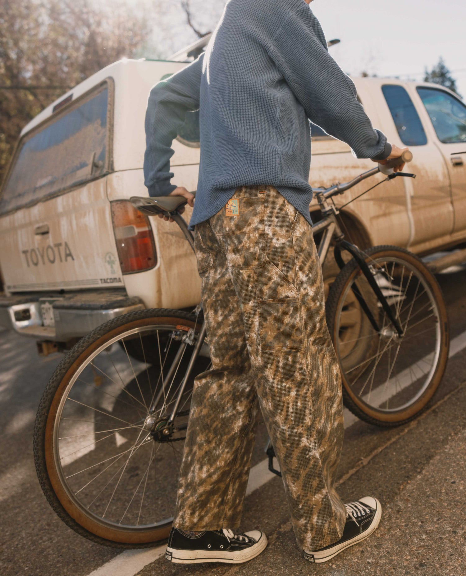 Person wearing camouflage pants and a blue shirt, standing next to a bicycle with a Toyota truck in the background.
