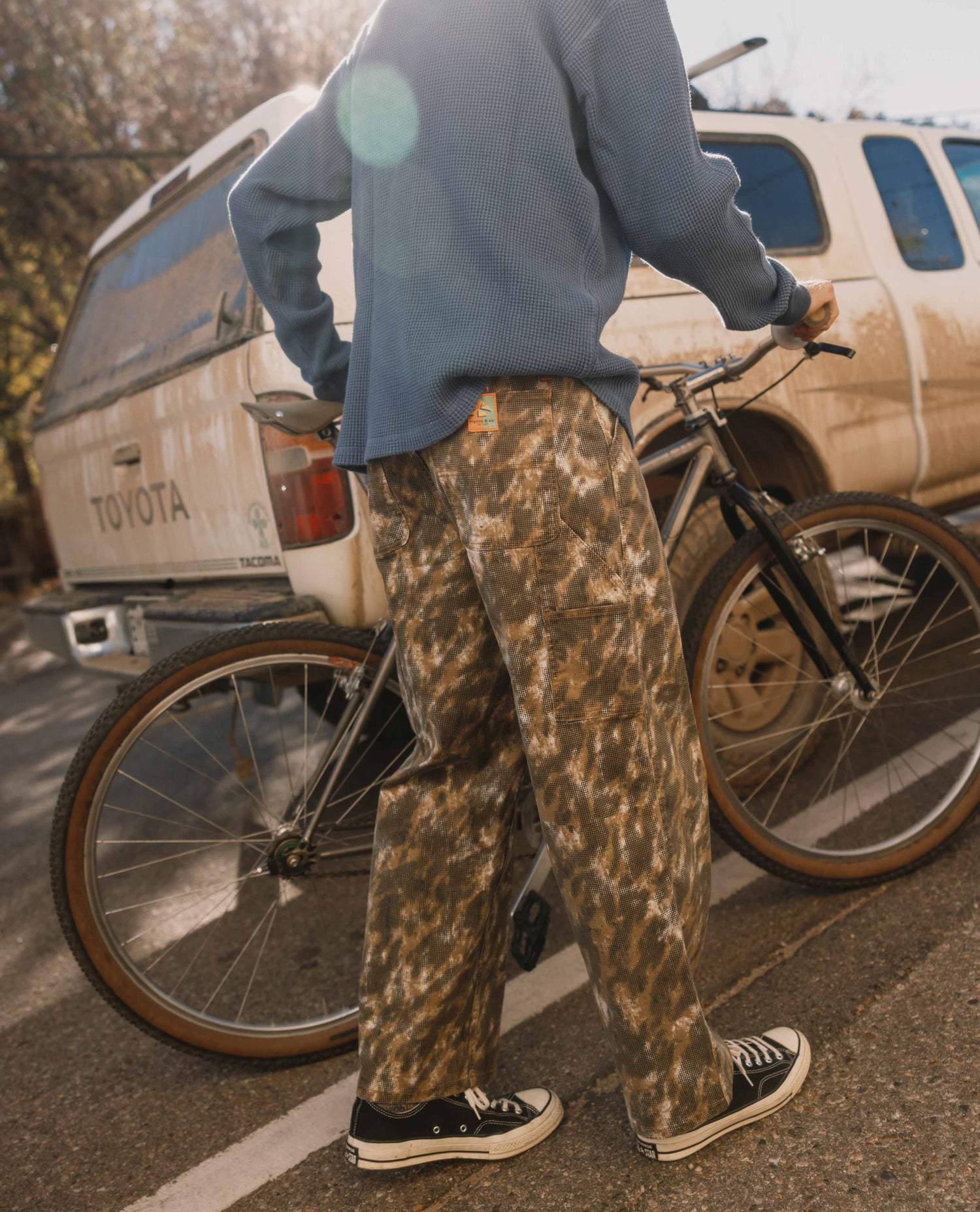 Person wearing camouflage pants and a blue jacket standing next to a bicycle with a Toyota van in the background.