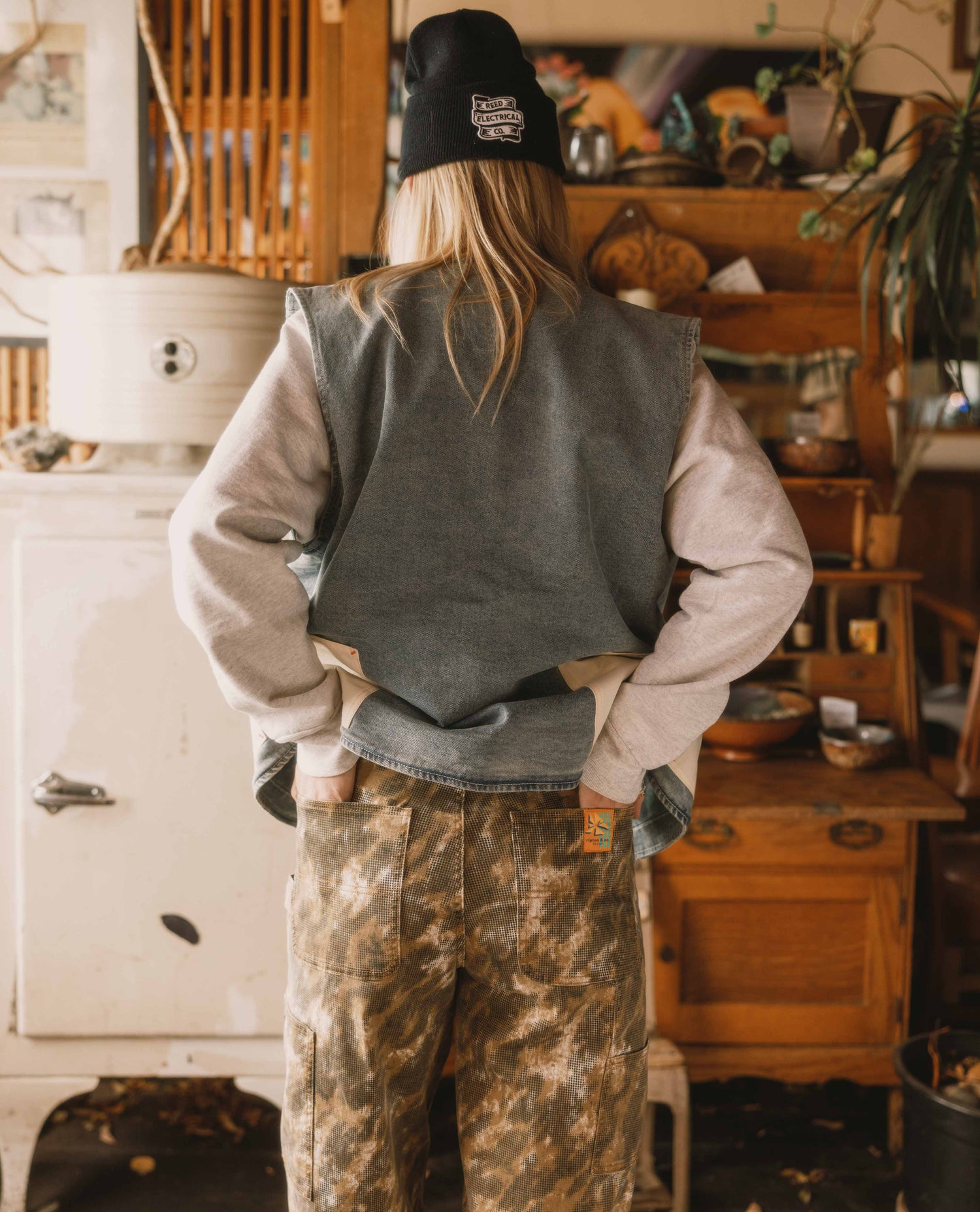 Person wearing a gray vest, beige sweater, and camouflage pants in a rustic indoor setting.