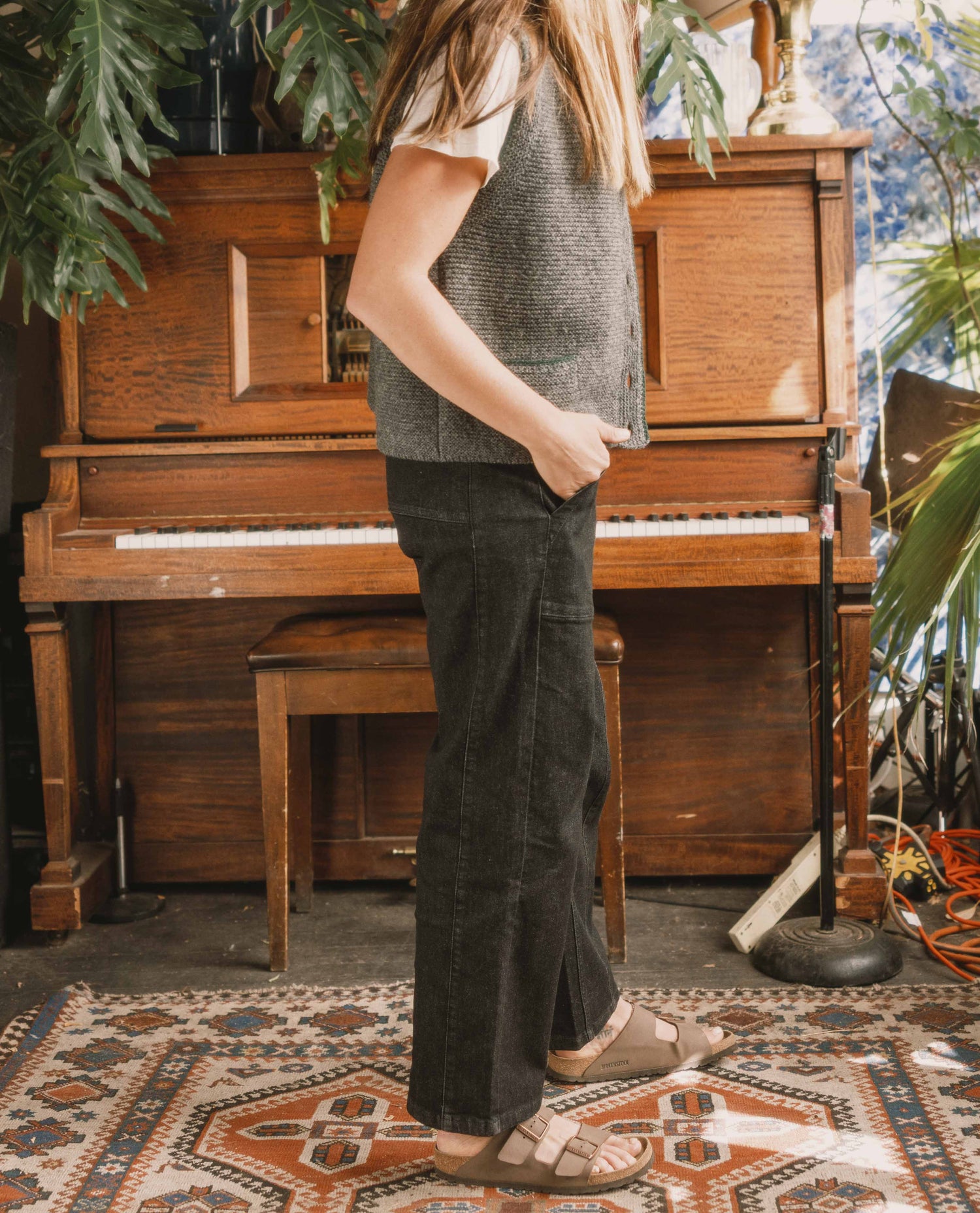 Person standing in front of a wooden piano in a room with plants and a patterned rug.