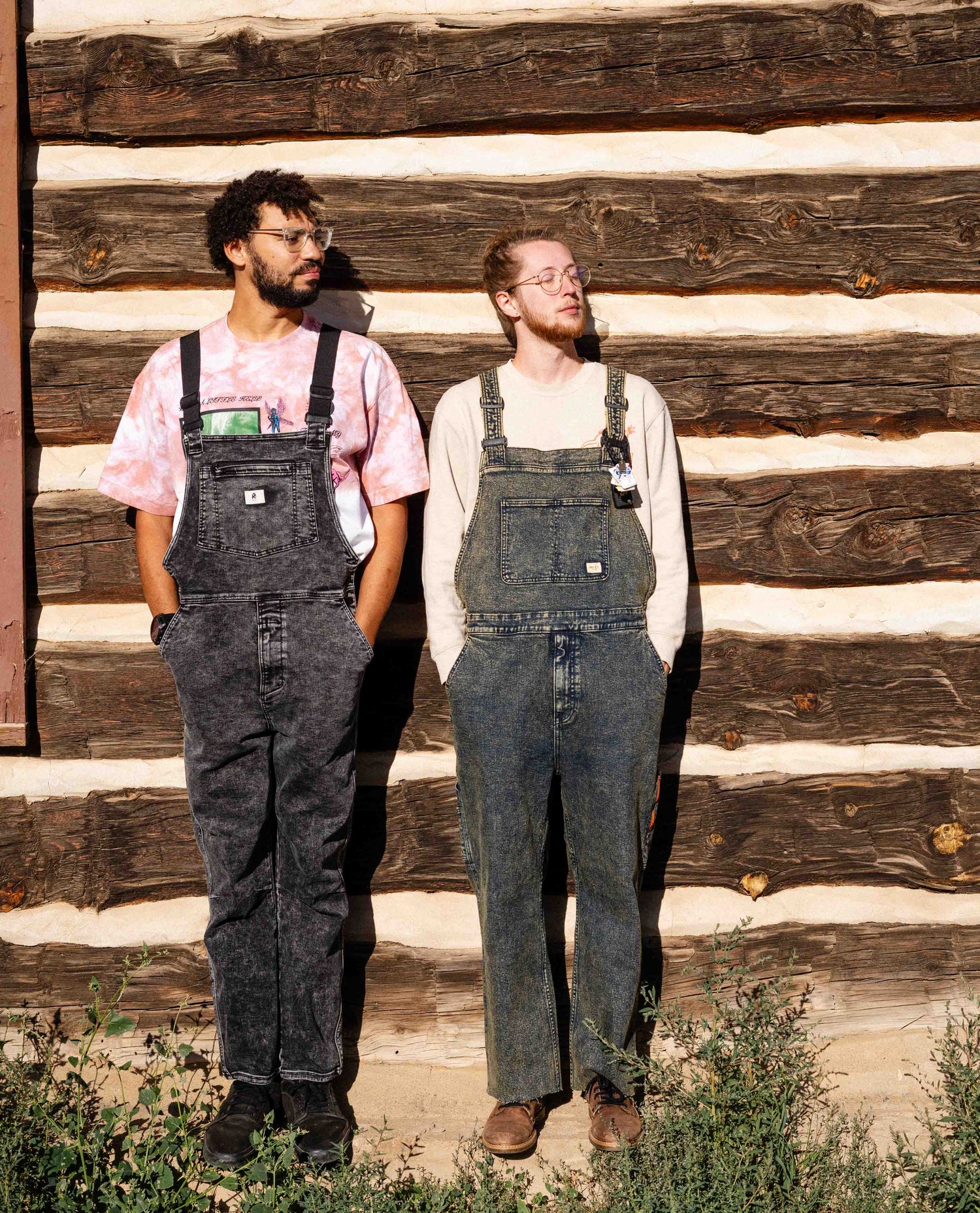 Two guys wearing overalls next to a log cabin