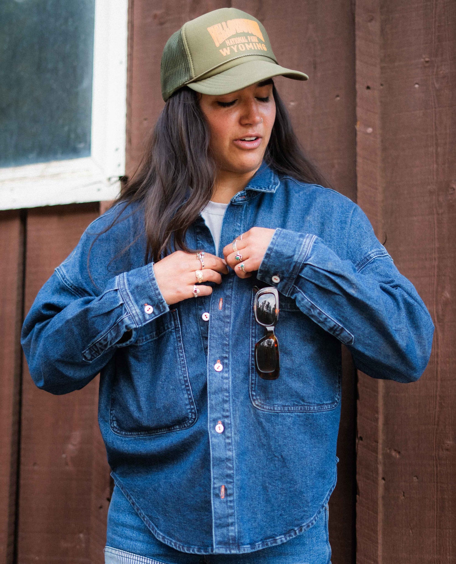 Person wearing a blue denim shirt and green cap with text, standing against a wooden wall.