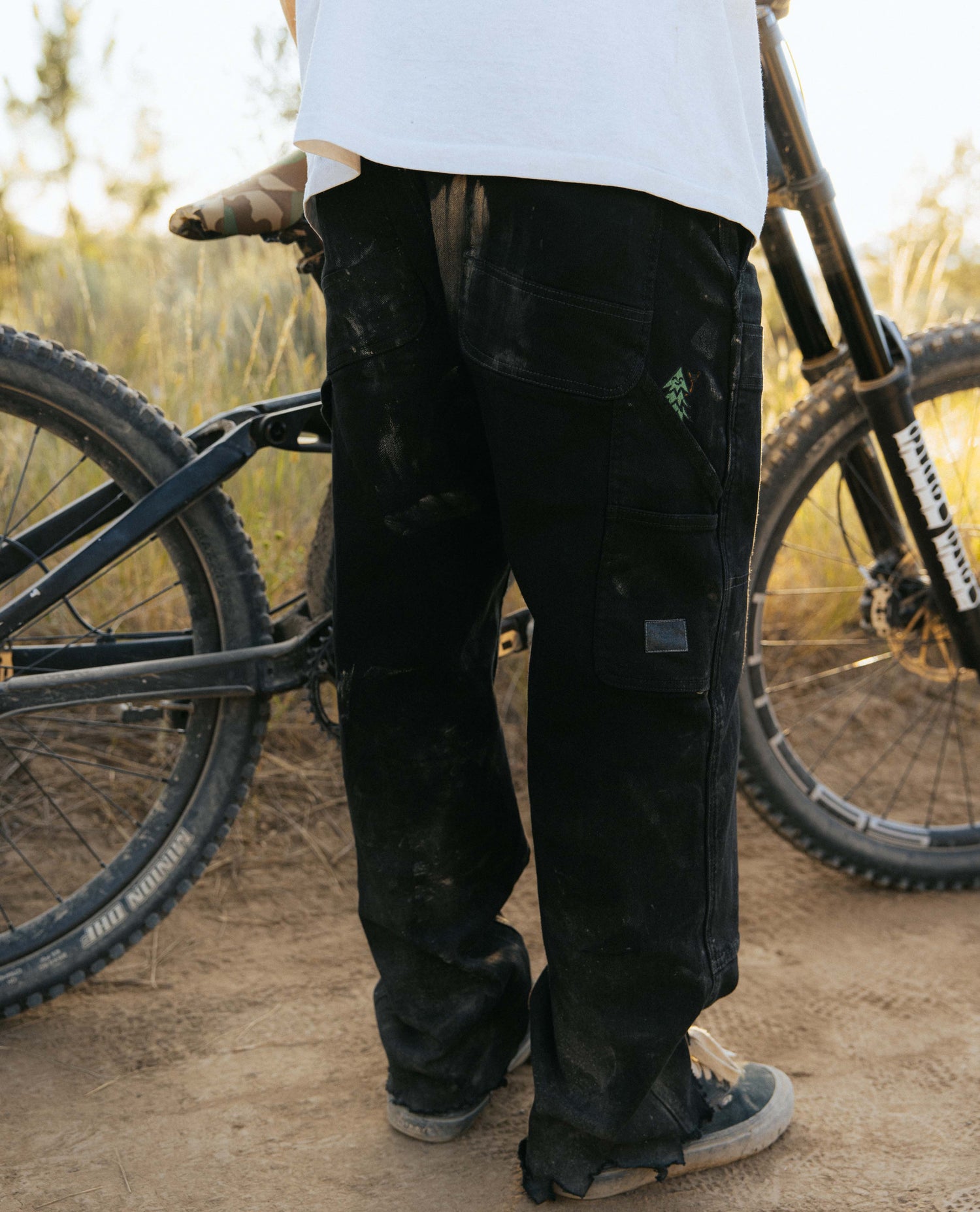 Person wearing black pants with a logo, standing next to a bicycle in an outdoor setting.