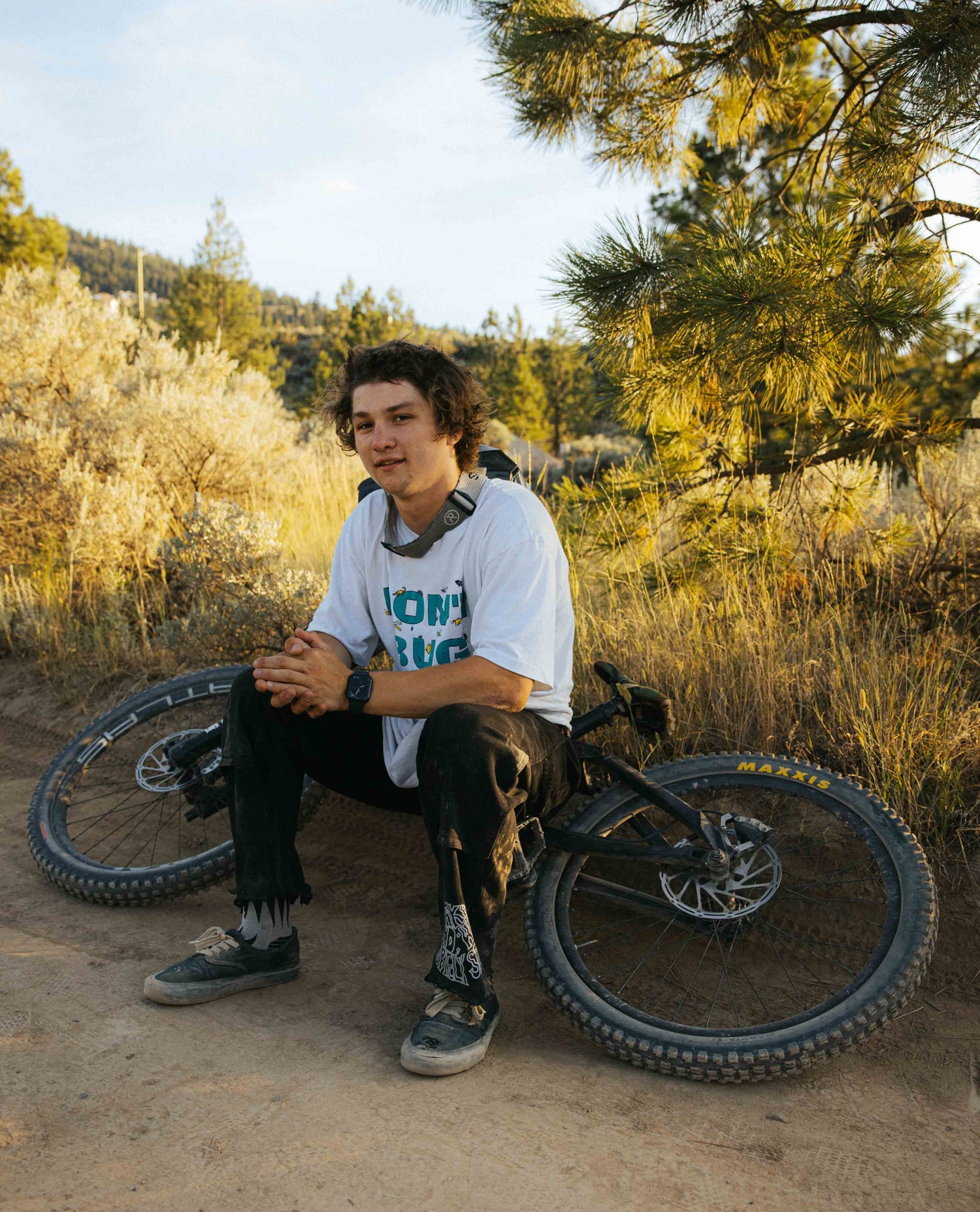 Person sitting on a dirt path with a bicycle, surrounded by trees and shrubs.