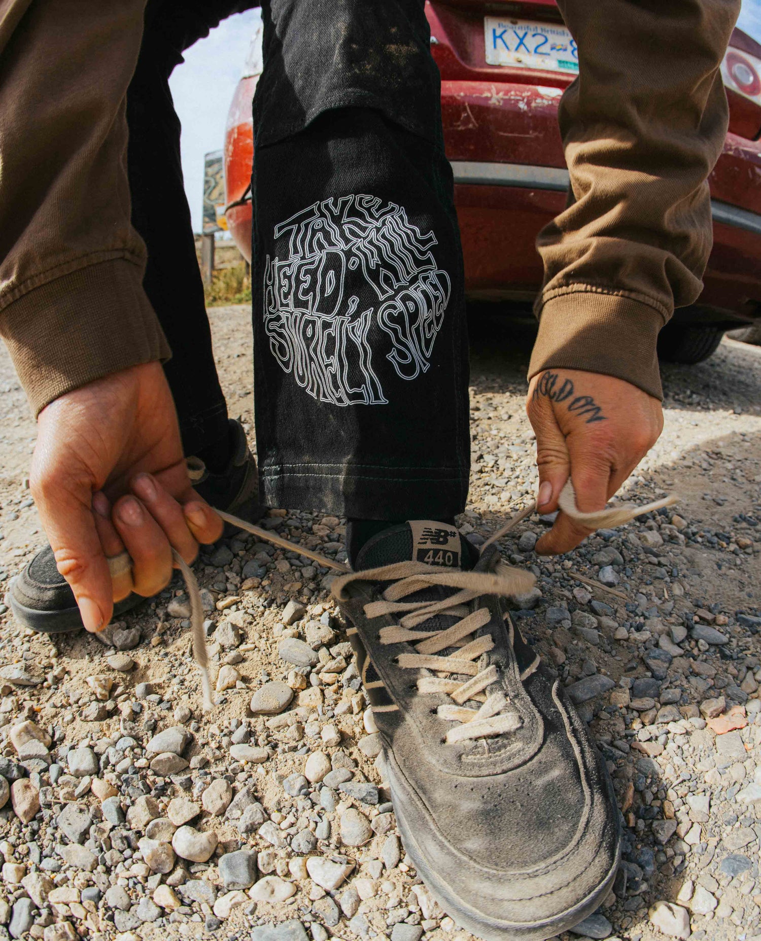 Person tying shoelaces on a pair of black sneakers with a visible brand logo, standing on a gravel surface.