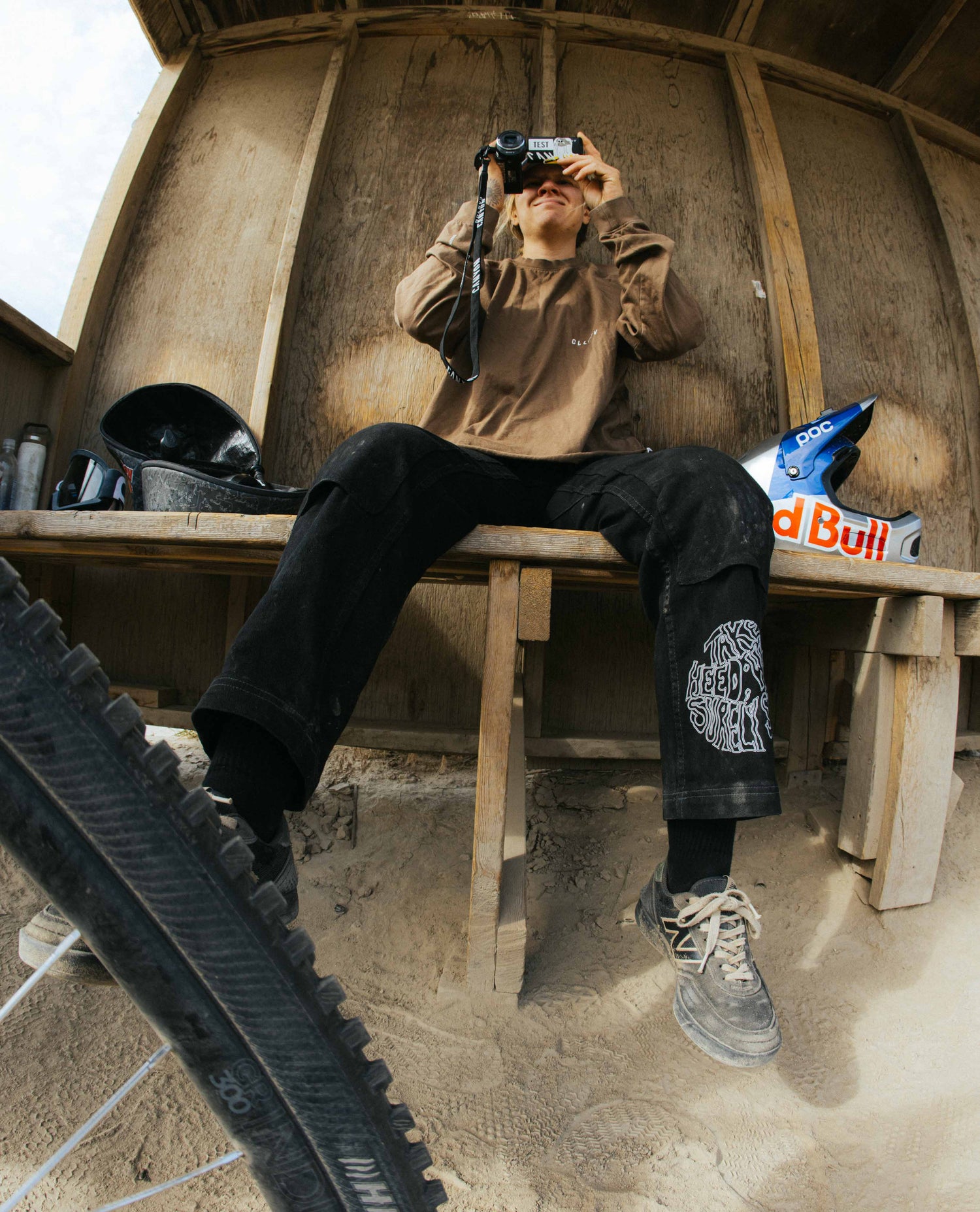 Person sitting on a wooden bench taking a photo with a camera, wearing a brown jacket and black pants.