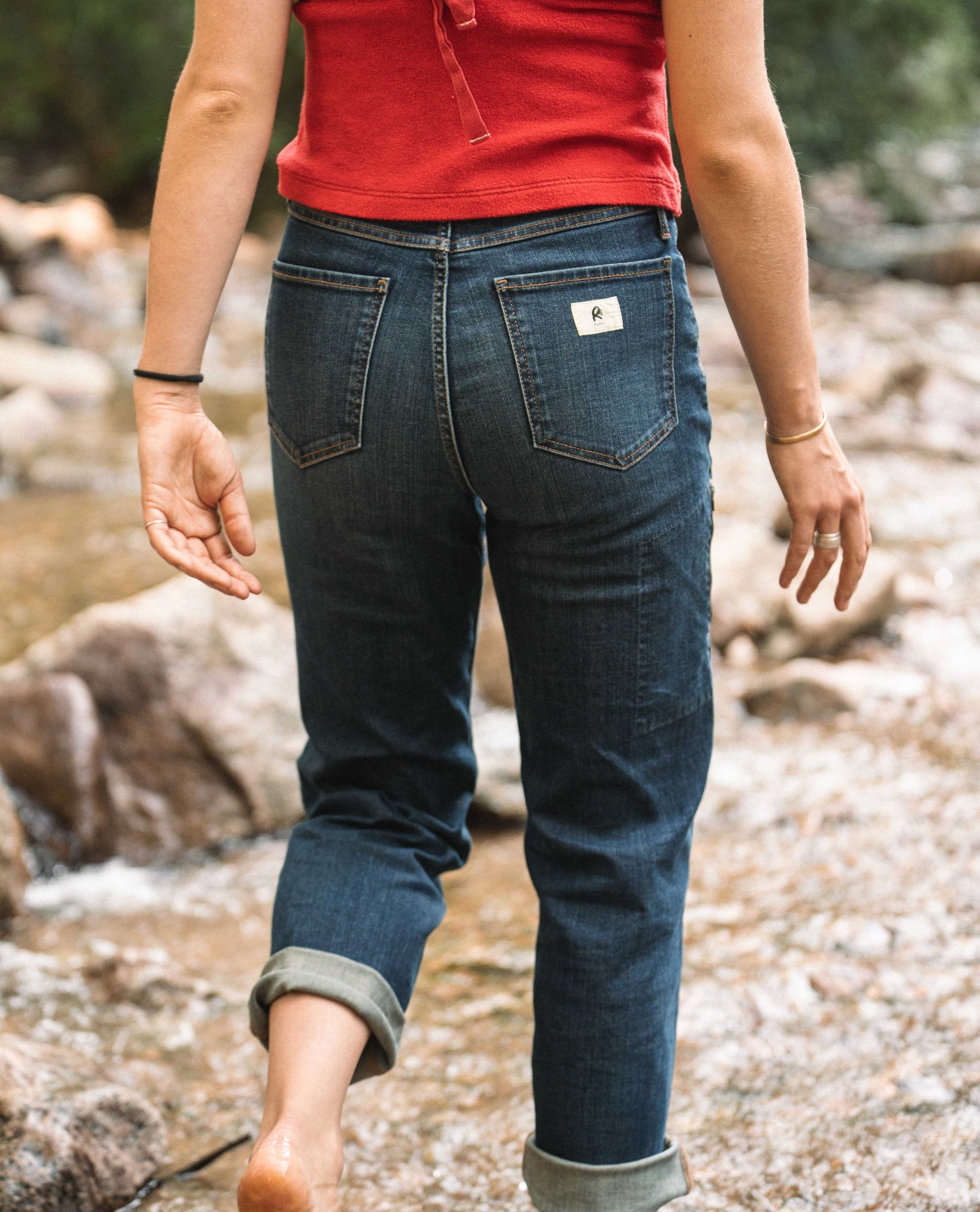 A woman walks away from camera through a shallow stream. She is wearing a red shirt and blue jeans.