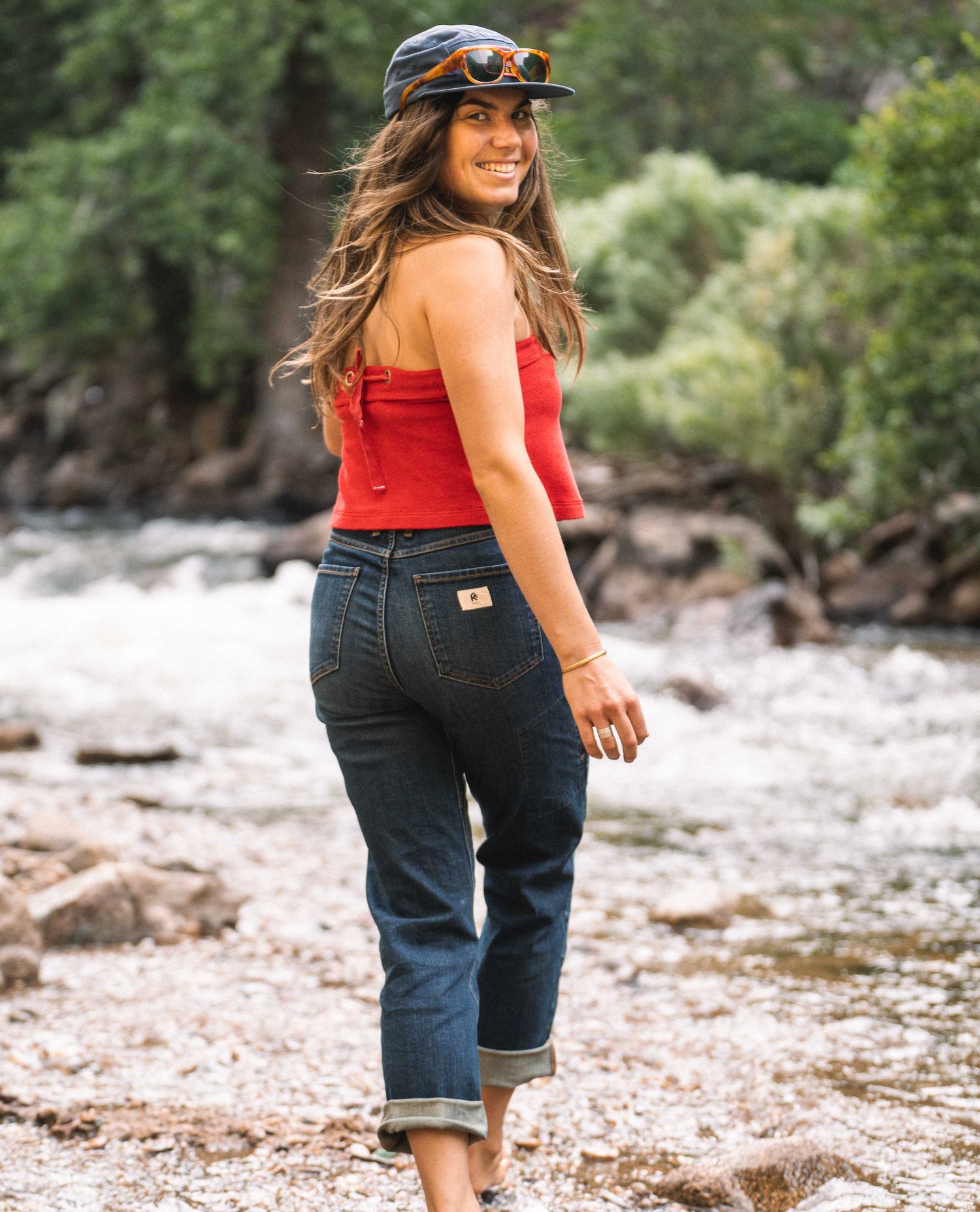 A woman walks away from and looks back at camera. She is walking through a shallow river. She is wearing a blue hat with glasses on the brim, a red tank top, and blue jeans.