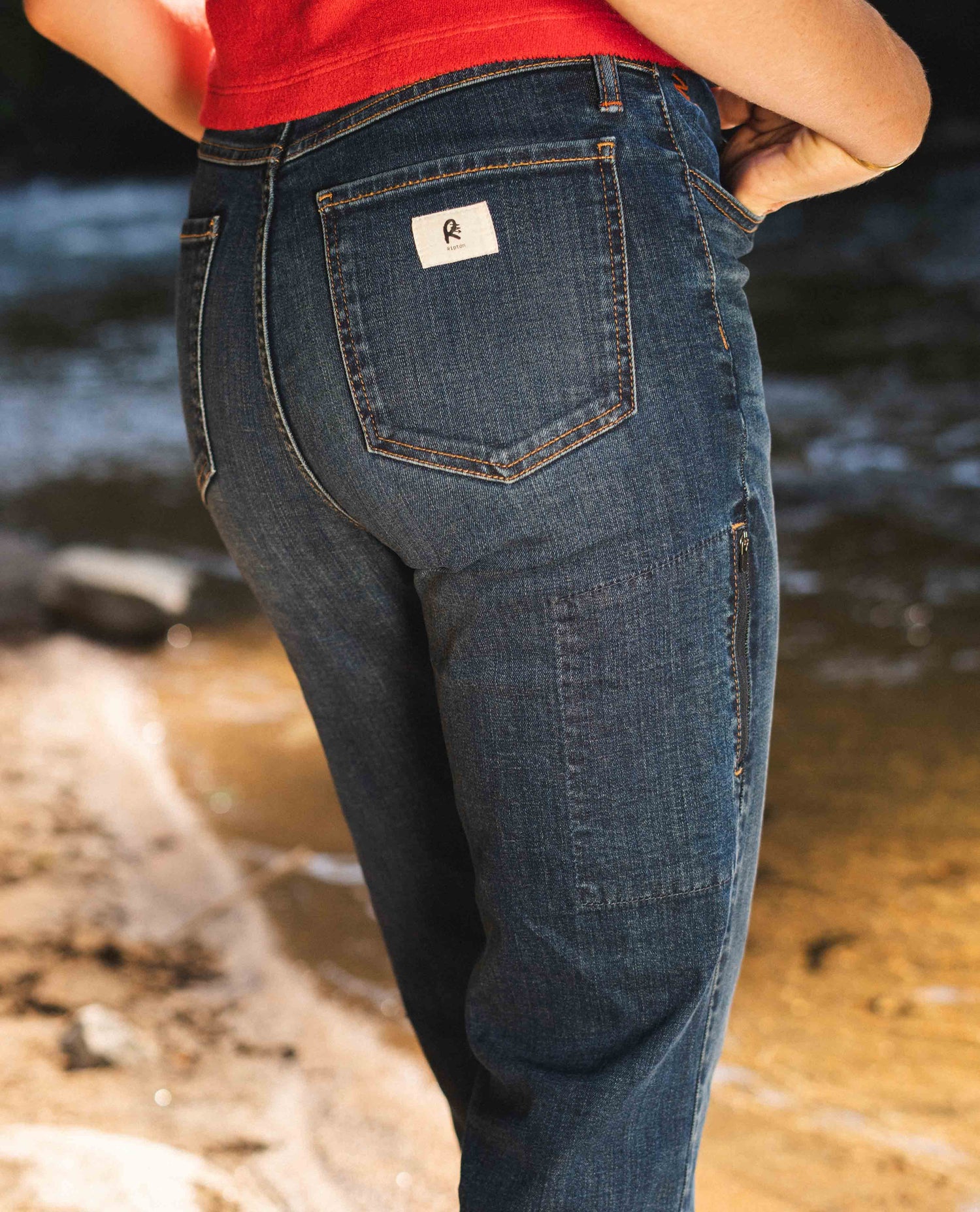 A rear view of a pair of Ripton blue jeans. Model is wearing a red shirt and posing in front of a river. There is a white "Ripton" logo on the right rear pocket of the pants.