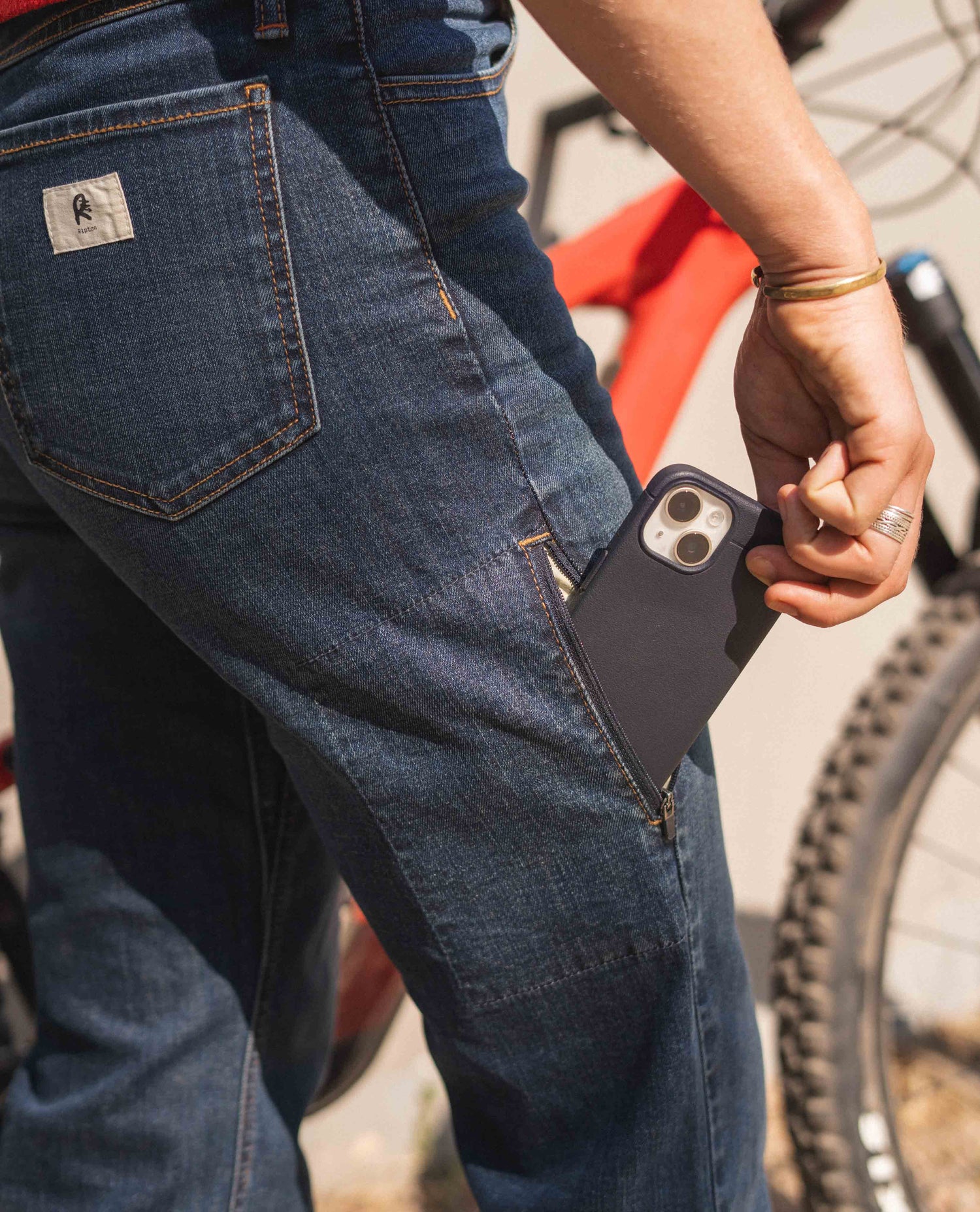 A woman pulling her phone out of a side pocket of a pair of blue jeans. There is an out of focused red and black mountain bike behind her.