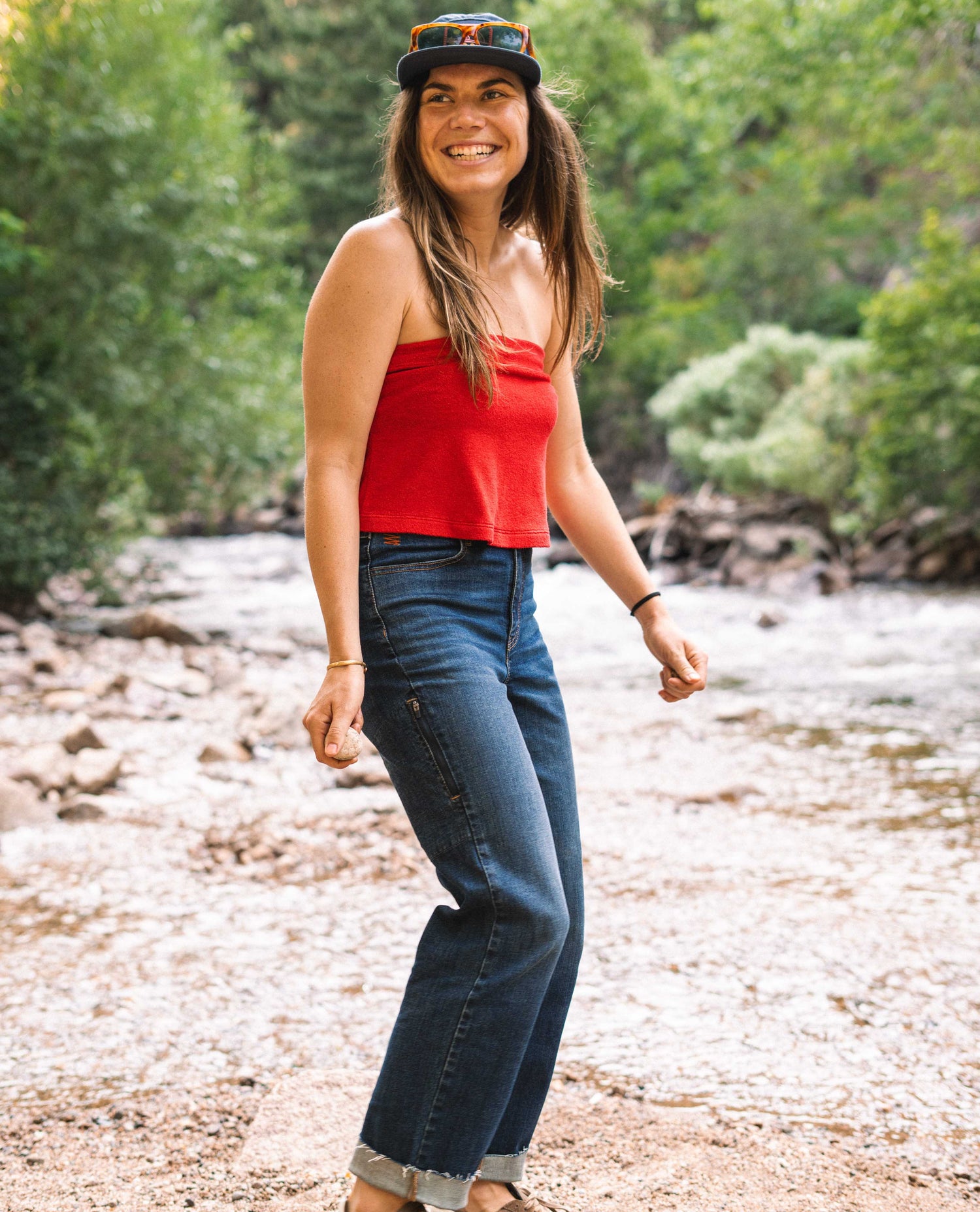 A woman standing on the shore of a stream. She is wearing a blue hat, a red tank top and dark blue jeans.