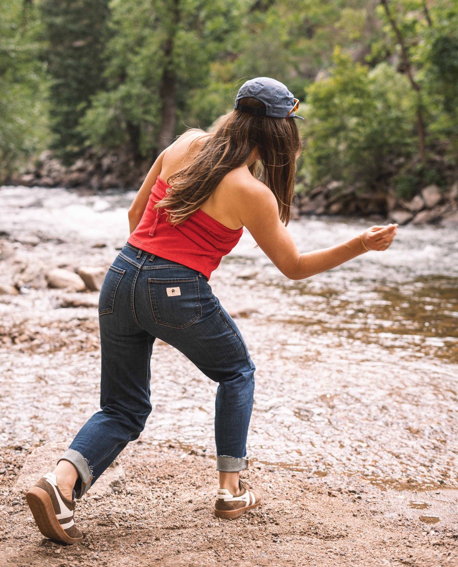 A woman skipping a rock on the side of a river. She wears a blue hat, a red shirt and blue jeans with brown shoes.