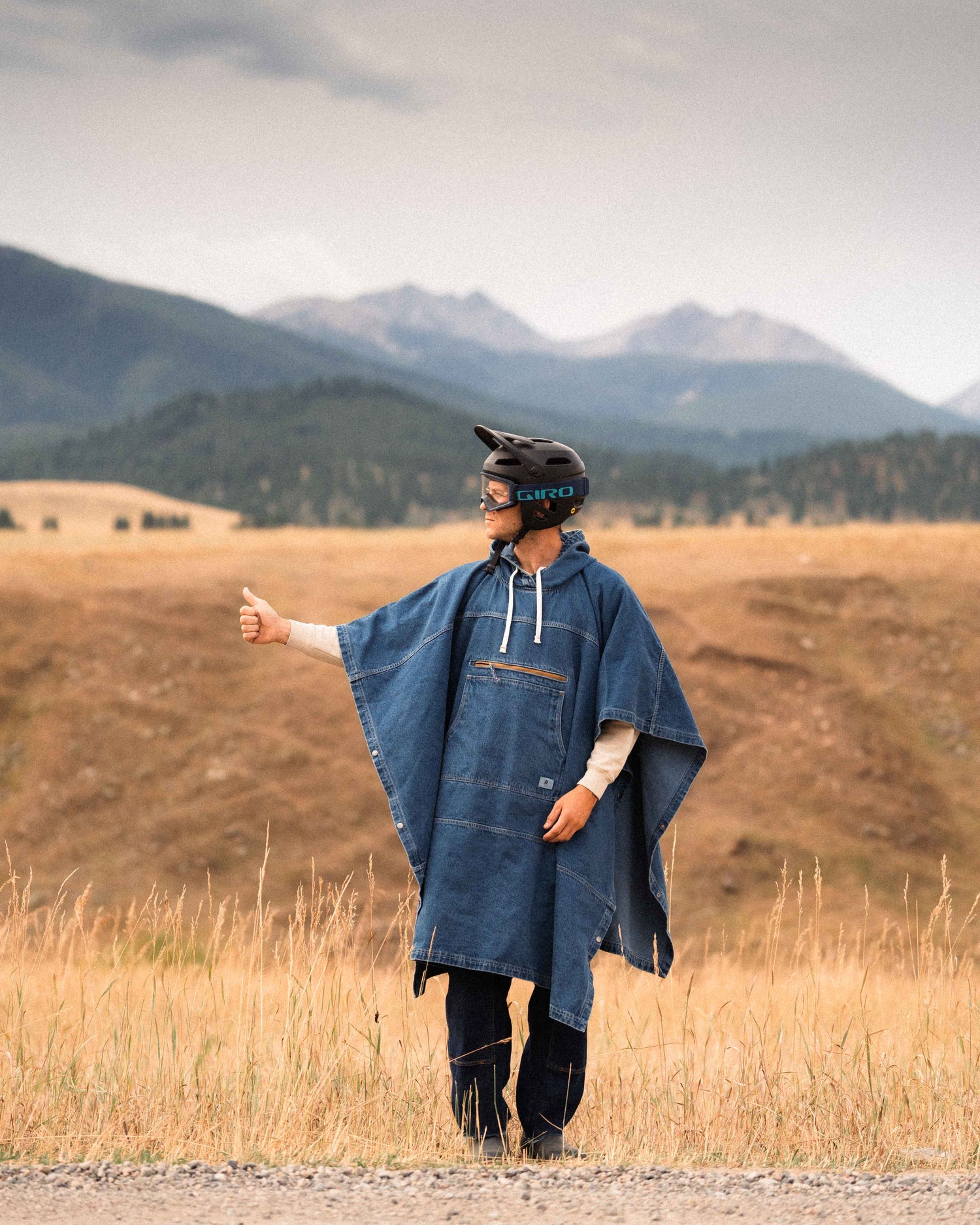 Person in blue poncho and helmet standing in a field with mountains in the background.