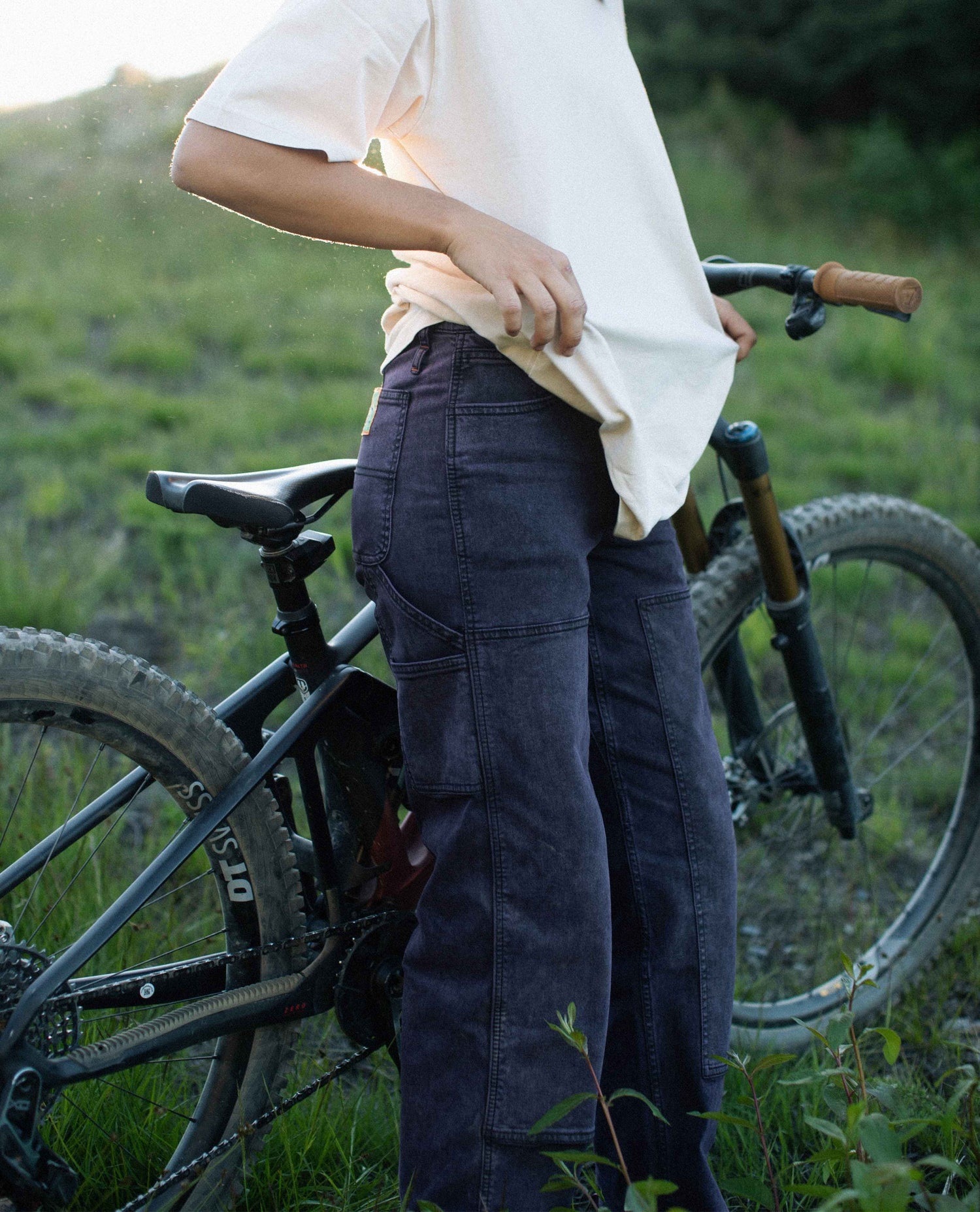 Person wearing dark pants and a light shirt standing next to a bicycle in a grassy outdoor setting