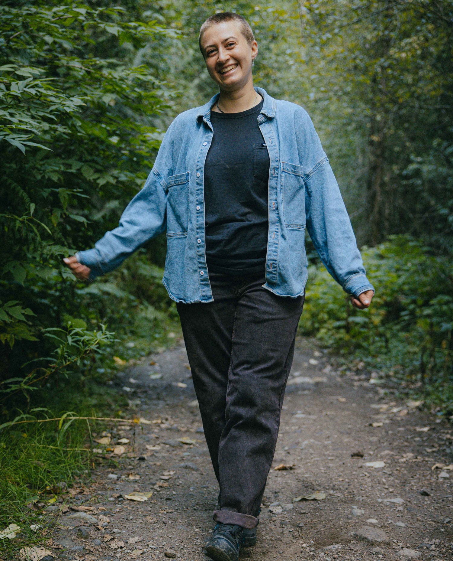 Person walking on a forest path wearing a denim shirt and dark pants.
