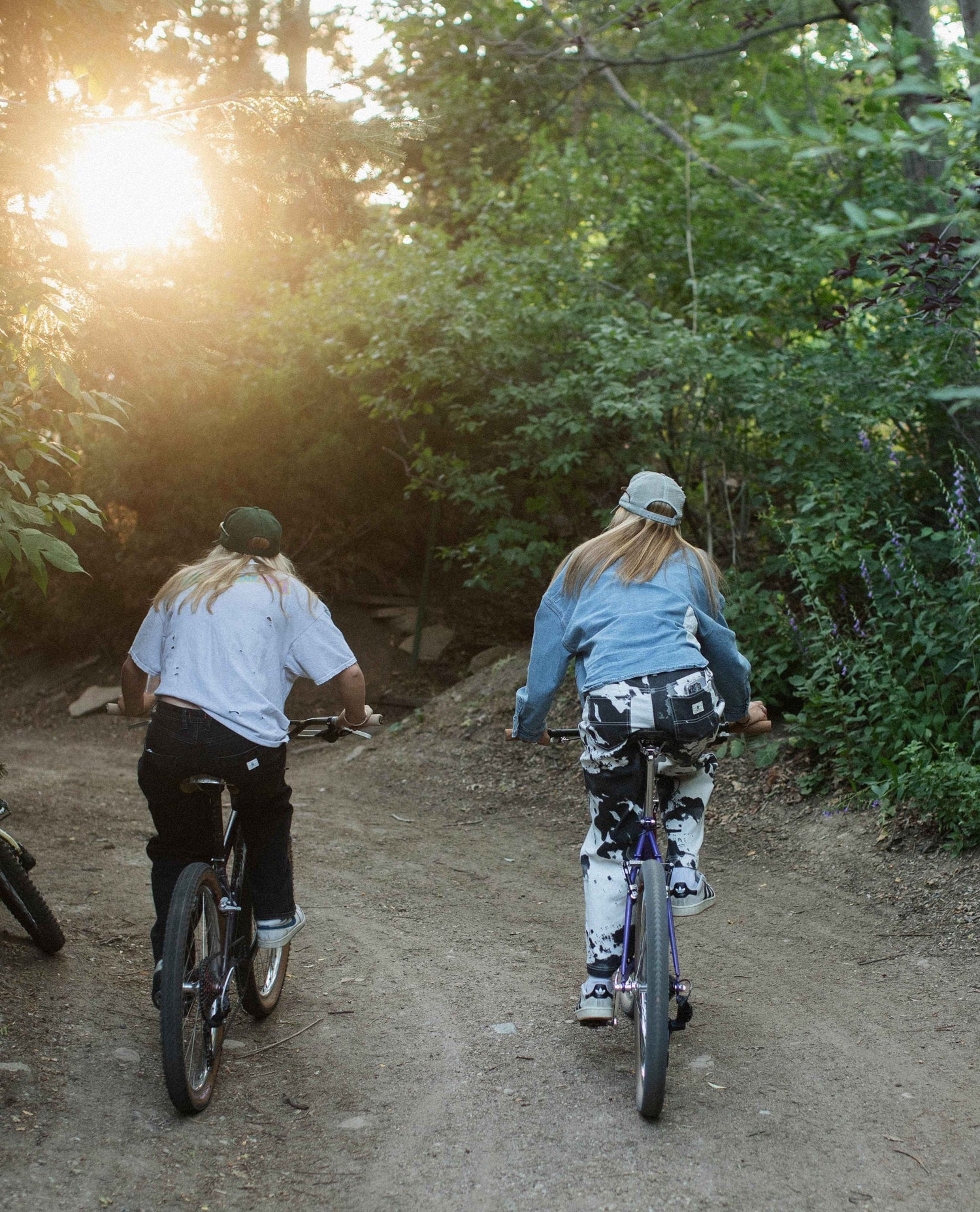 Two people riding bicycles on a dirt path through a forest with the sun shining through the trees.