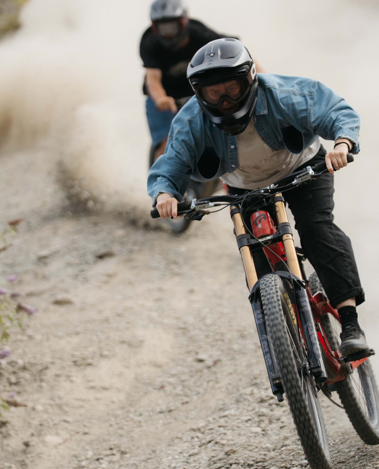 Two people riding bicycles on a dirt path with one person in the foreground wearing a helmet and goggles.