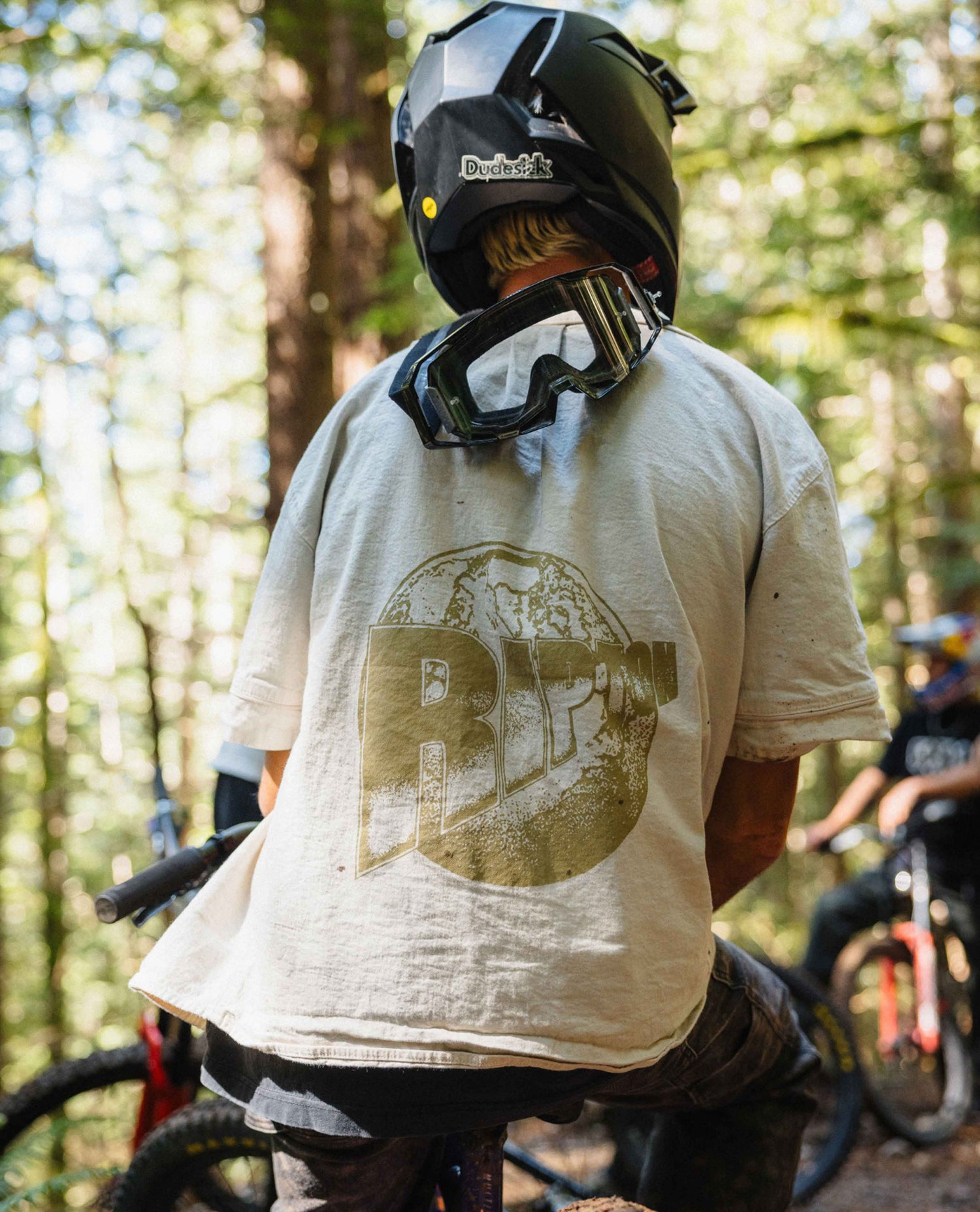A person on a mountain bike in a forest setting, viewed from behind. They are wearing a black helmet with goggles resting on their neck and a light beige graphic t-shirt with a large distressed brown circular logo on the back.