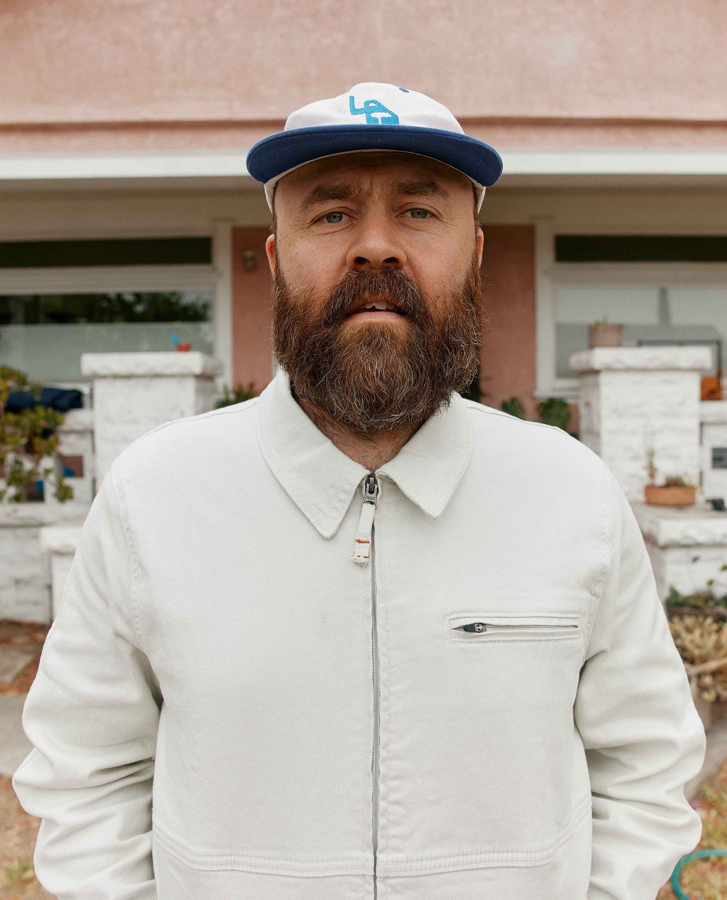 Man wearing white sport utility jacket with a white hat in front of a house.