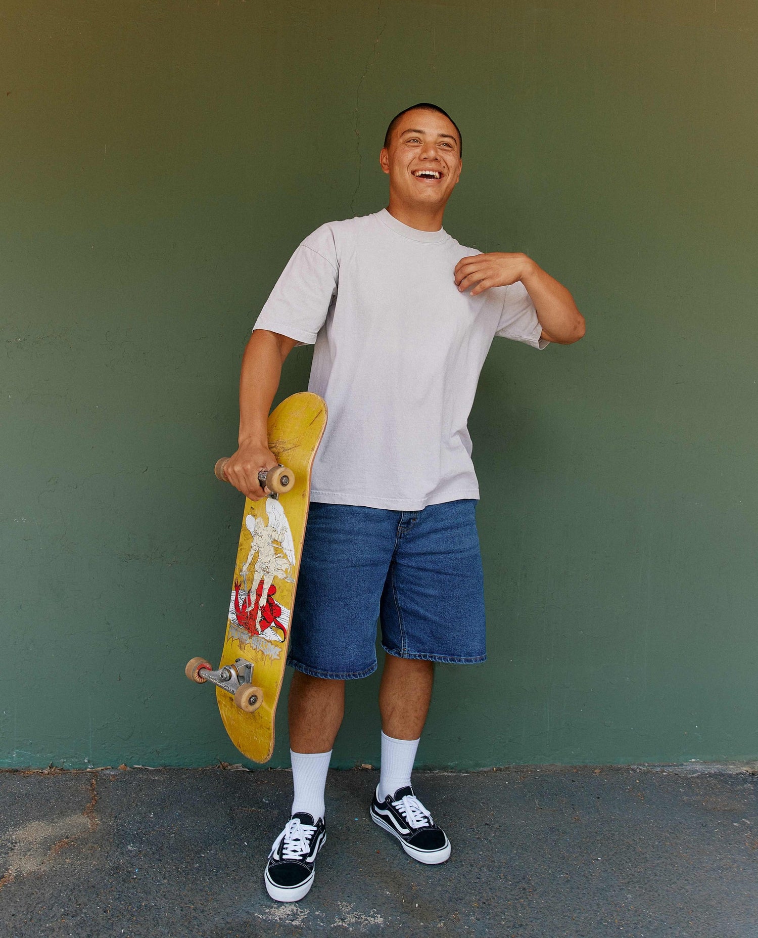 Man holding a skateboard wearing a white t shirt and blue jean shorts. 