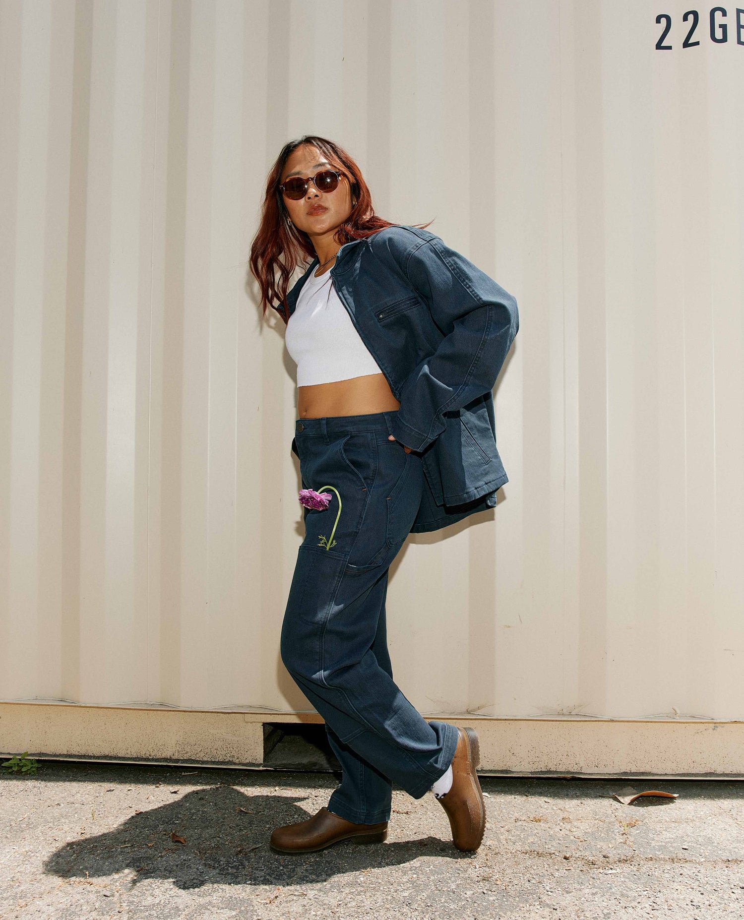 A woman in a white crop top, blue cargo style jacket, blue cargo pants, and round brown sunglasses posing in front of a cargo container with a pink flower in her left pocket. 