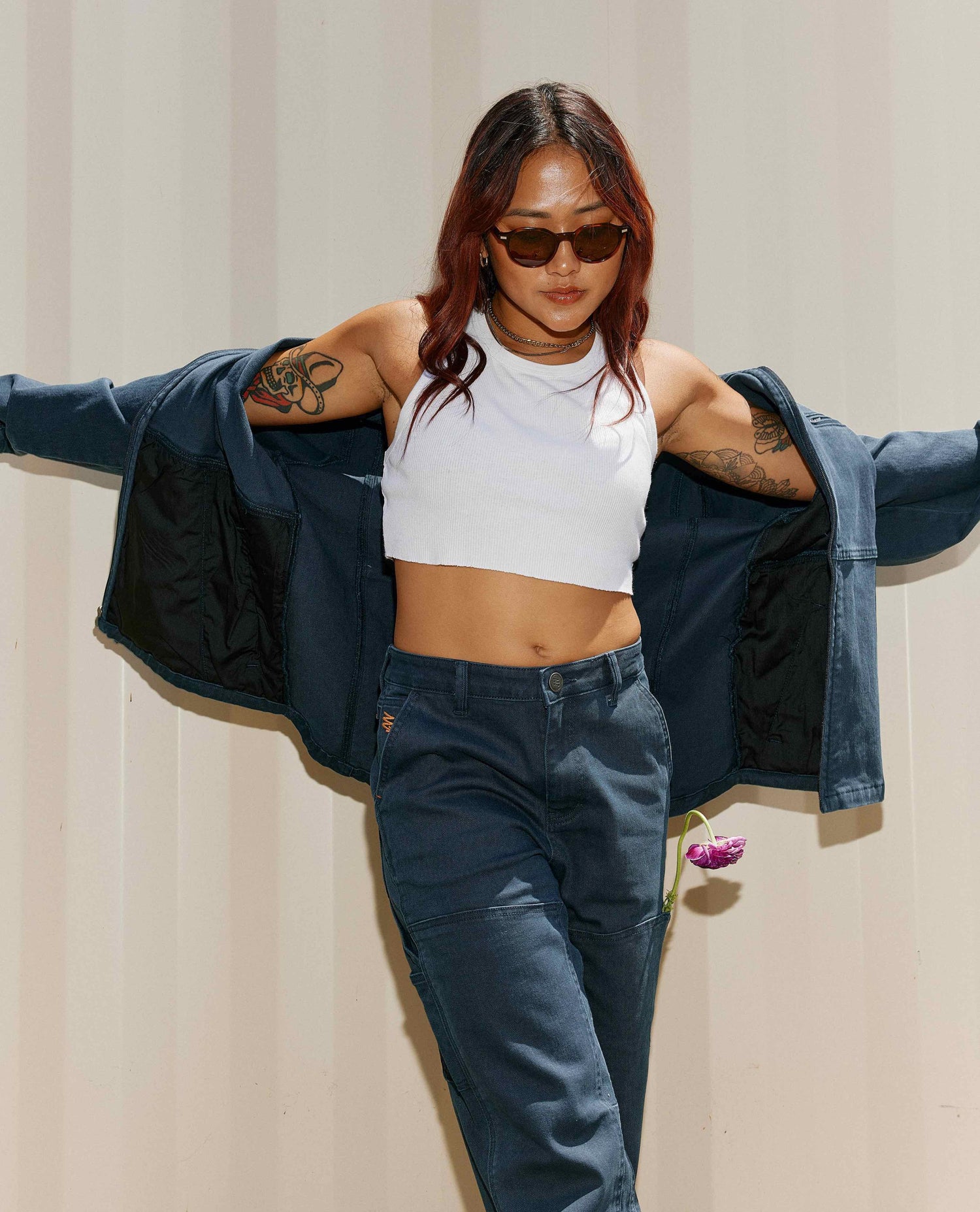 A woman in front of a tan cargo container in a white crop top with a matching set of blue cargo style jacket and pants. She has a pink flower in her left leg pocket. She is also wearing brown round sunglasses. 