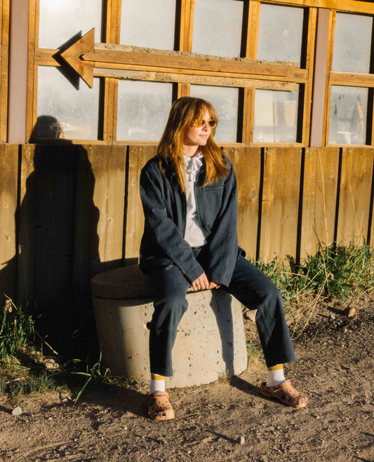 A woman sitting down on a cement cylinder looking off to her left. She is wearing a blue cargo style jacket and pants as well as a light grey hoodie and light brown sunglasses. She is seated in front of an old building. 