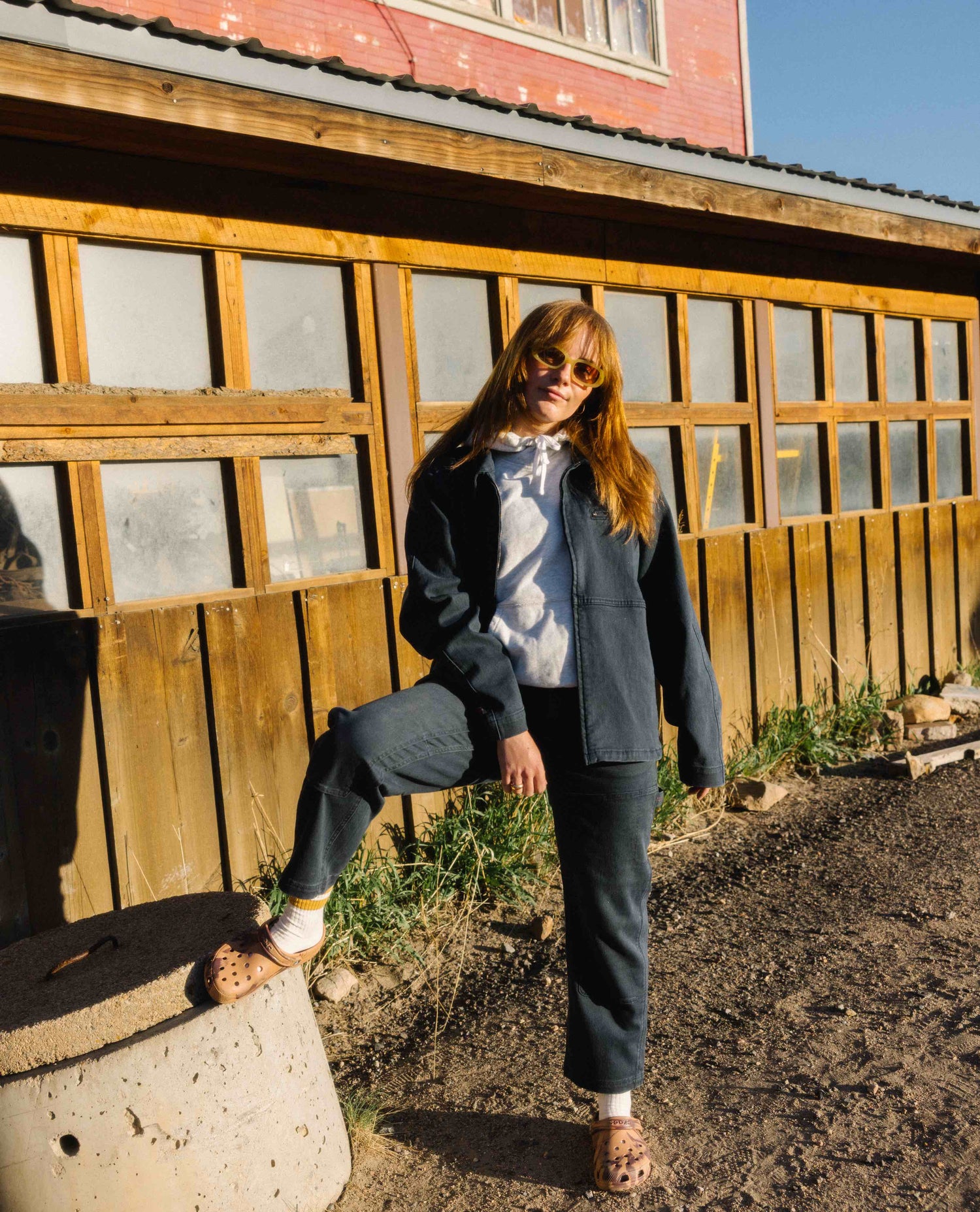 A woman in front of an old building wearing a blue cargo style jacket and pants with a grey hoodie, sunglasses and brown crocs. her right leg is posed on top of a cement cylinder. 