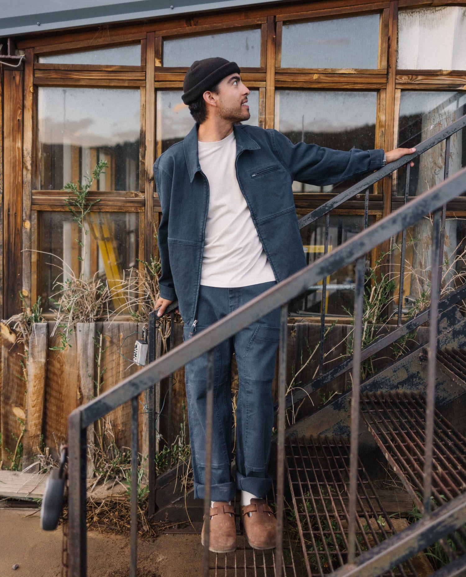 A man in a white t shirt and blue cargo style jacket and pants standing in front of an old window looking off to his left. He is also wearing a black beanie and brown shoes. 