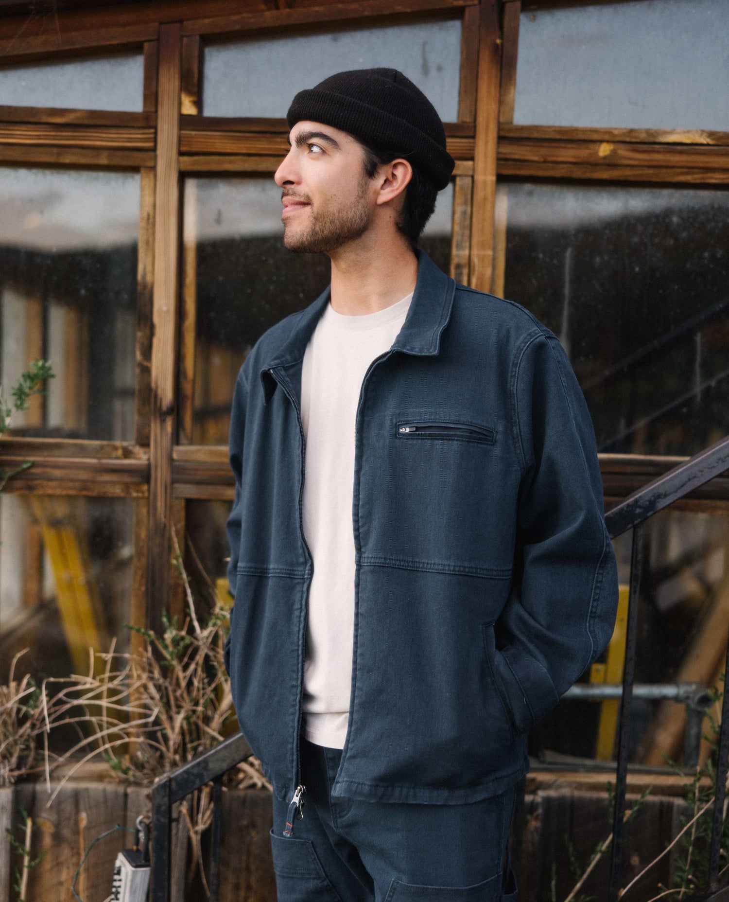 A man standing in front of an old buildings window with a black beanie on his head. He is wearing a white t shirt with a blue cargo style jacket and pants. He is looking off to his right. 