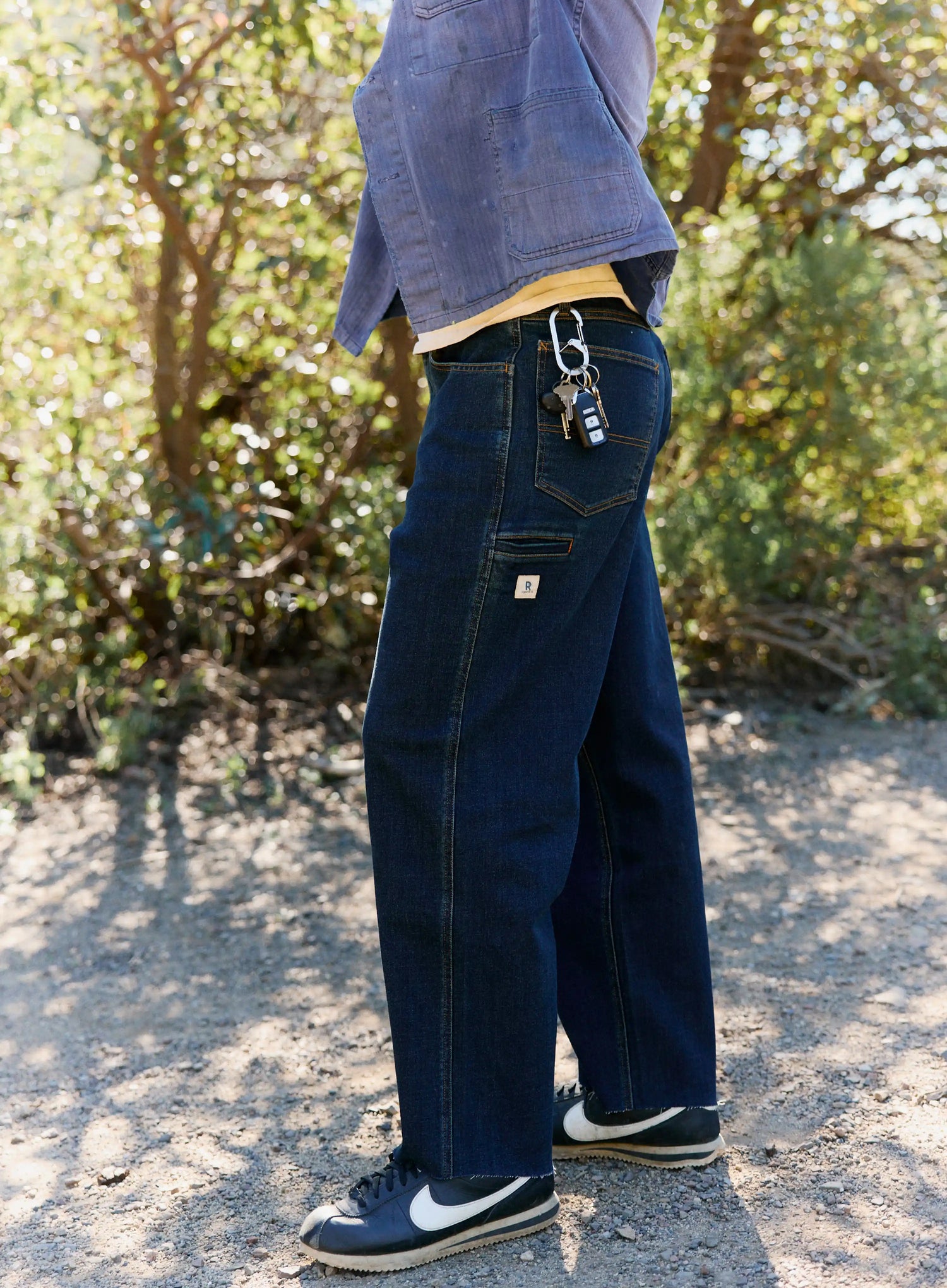 Person wearing blue jeans with a visible brand logo, standing outdoors with greenery in the background.
