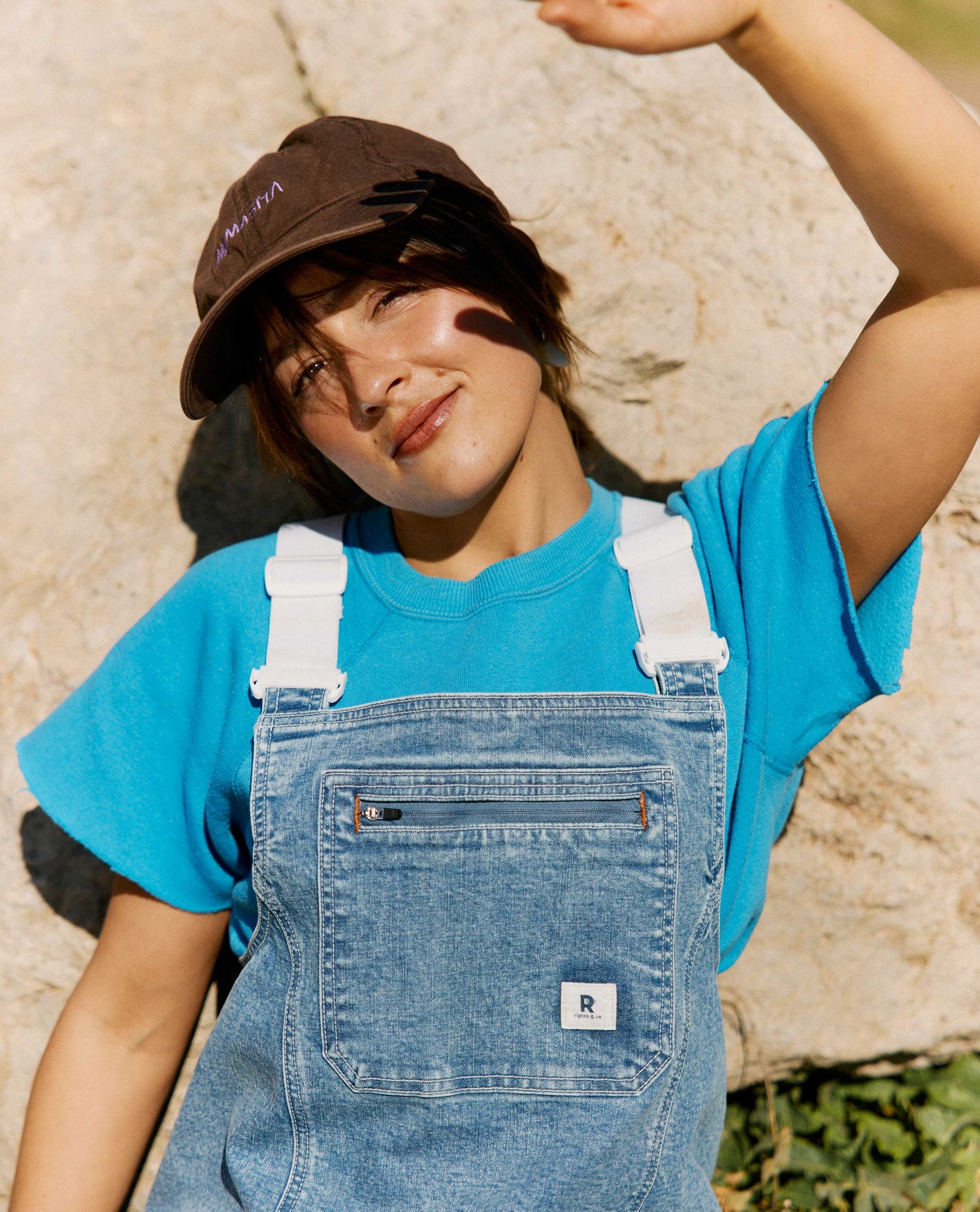 Person wearing a blue shirt and denim overalls with a brown cap outdoors.