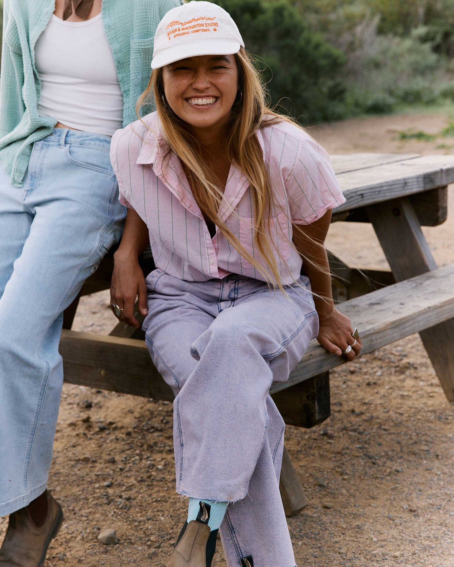 Two women sitting on a wooden bench outdoors, one wearing a white cap with text.