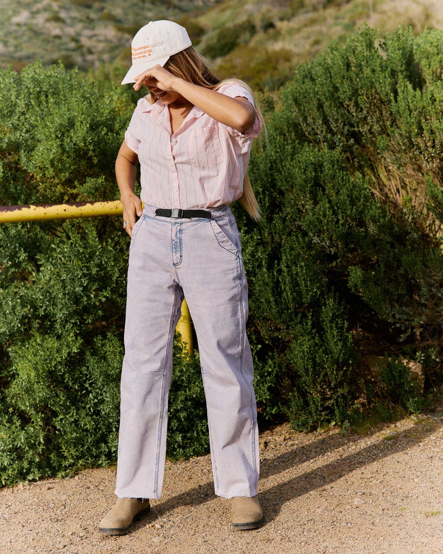 Person wearing a pink shirt and light-colored pants standing outdoors with greenery in the background