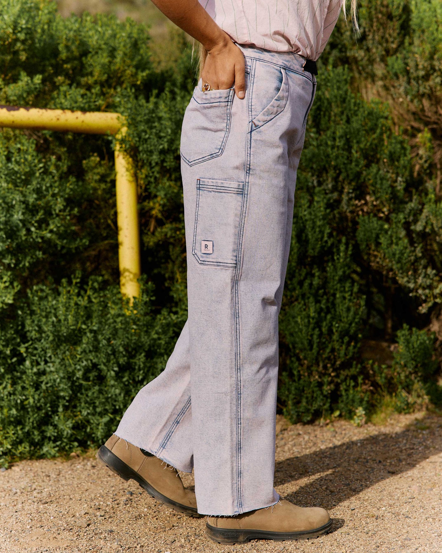 Person wearing light blue jeans with a brand logo, standing outdoors with greenery in the background.