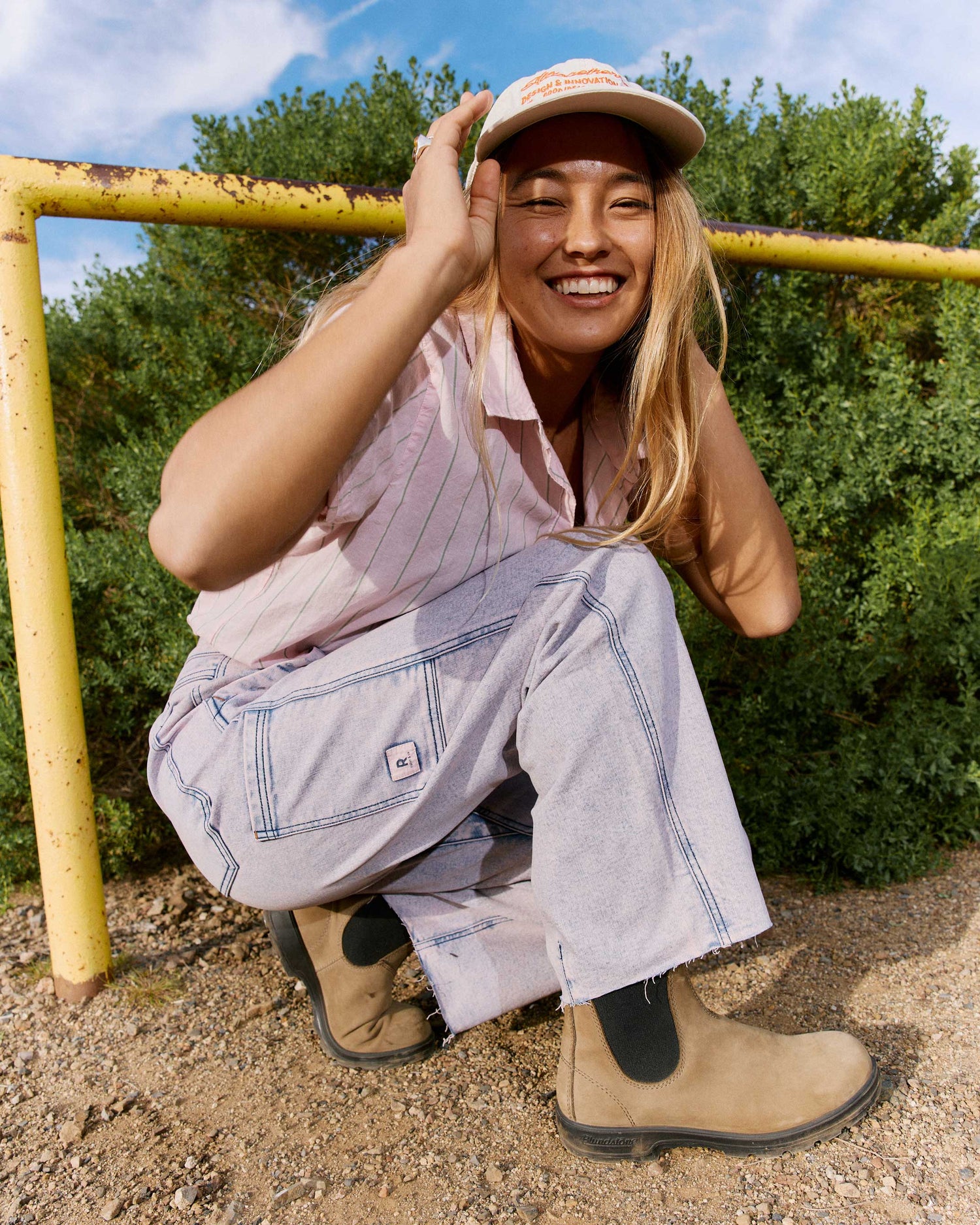 Person wearing a pink shirt, blue overalls, and tan boots sitting on a yellow metal structure with greenery in the background.