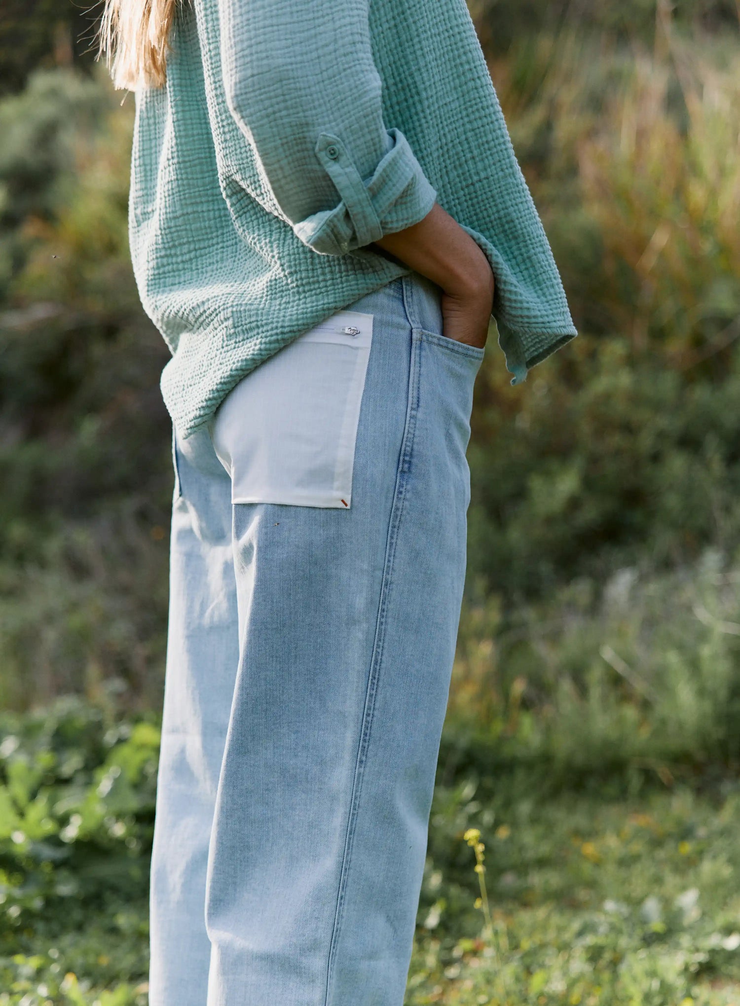 Person wearing a green sweater and light blue jeans standing in a natural setting.
