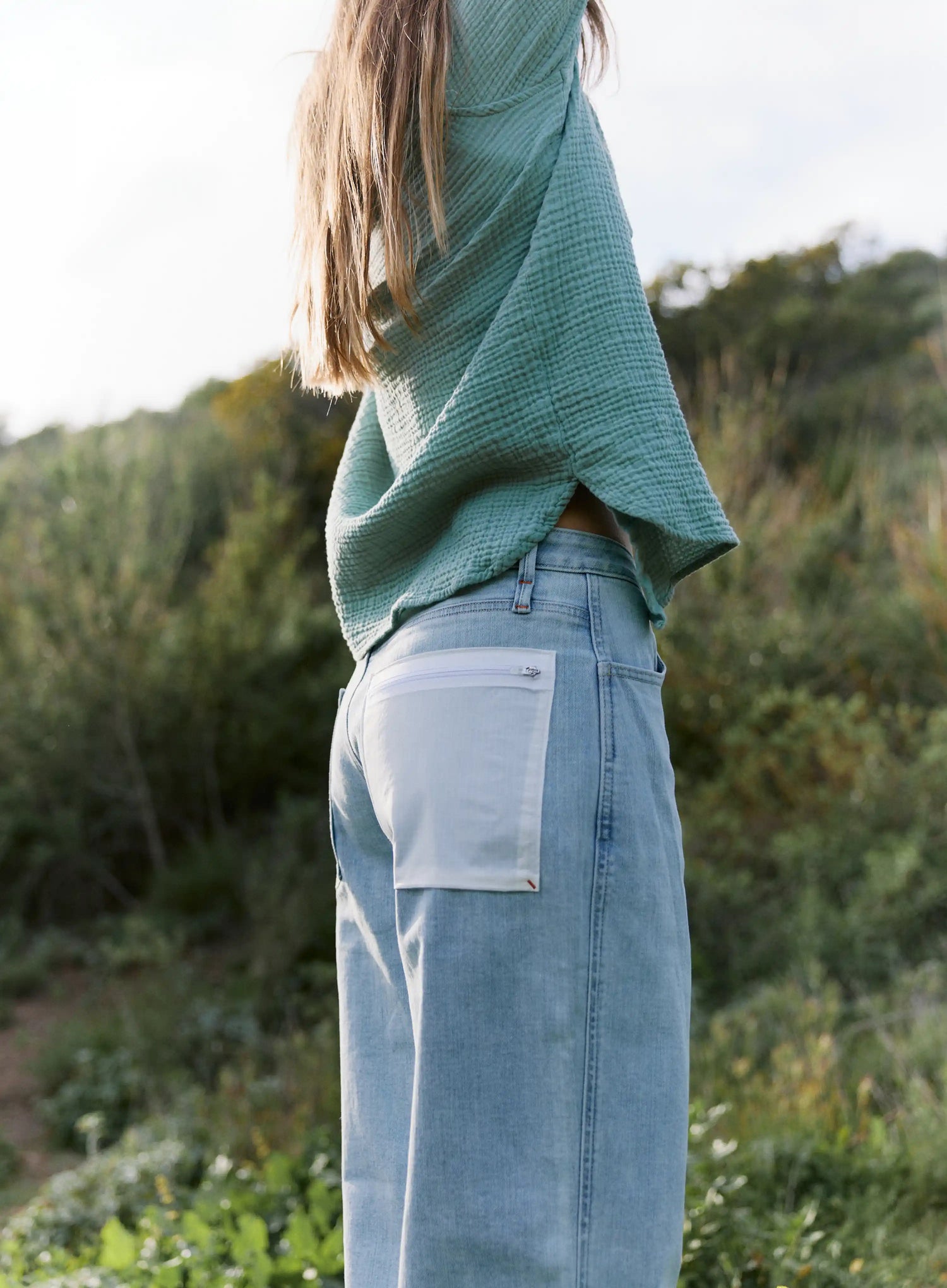 Person wearing a green top and light blue jeans standing in a natural setting with greenery.