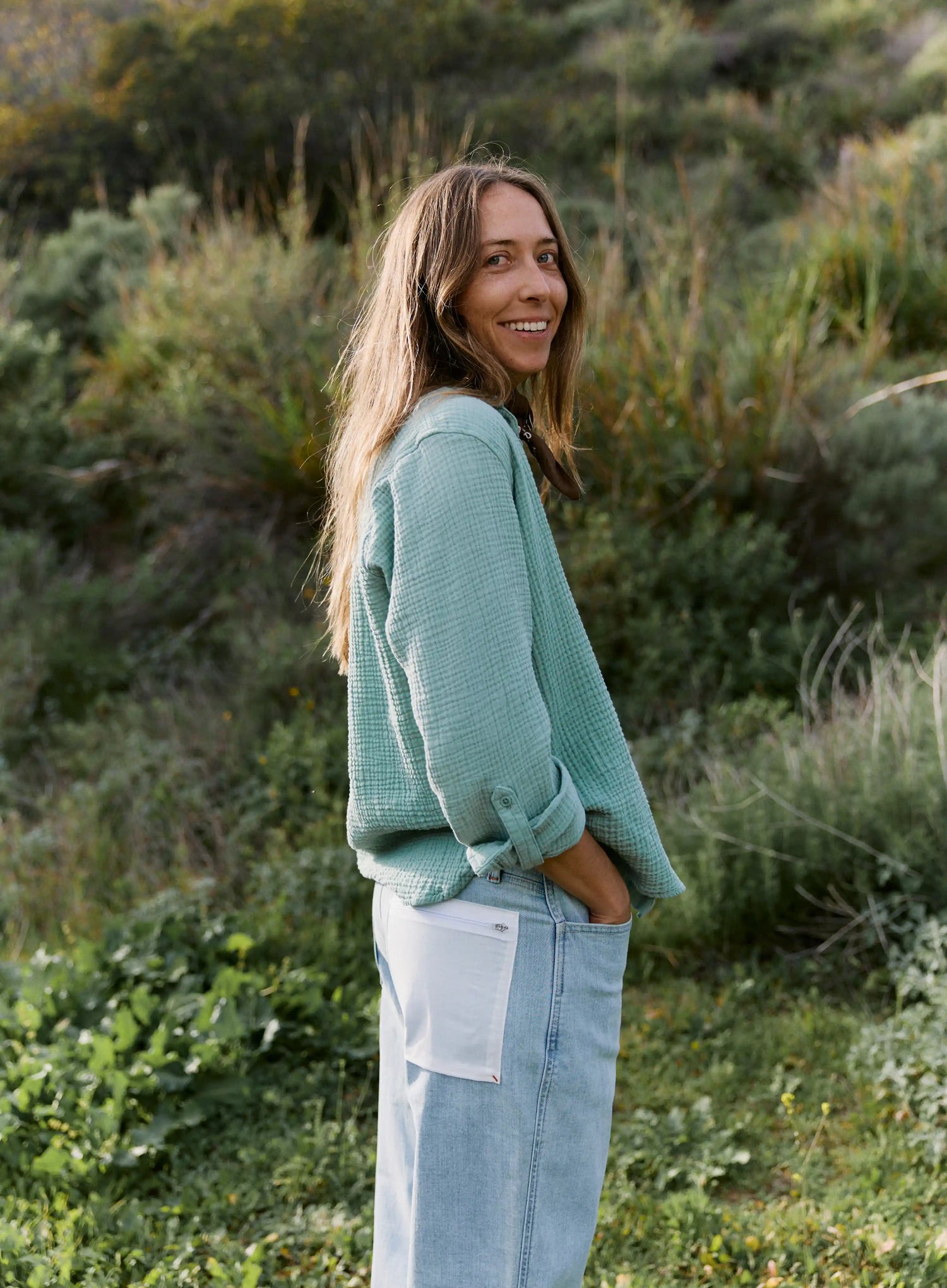 Woman in a green sweater and light blue jeans standing in a grassy field.