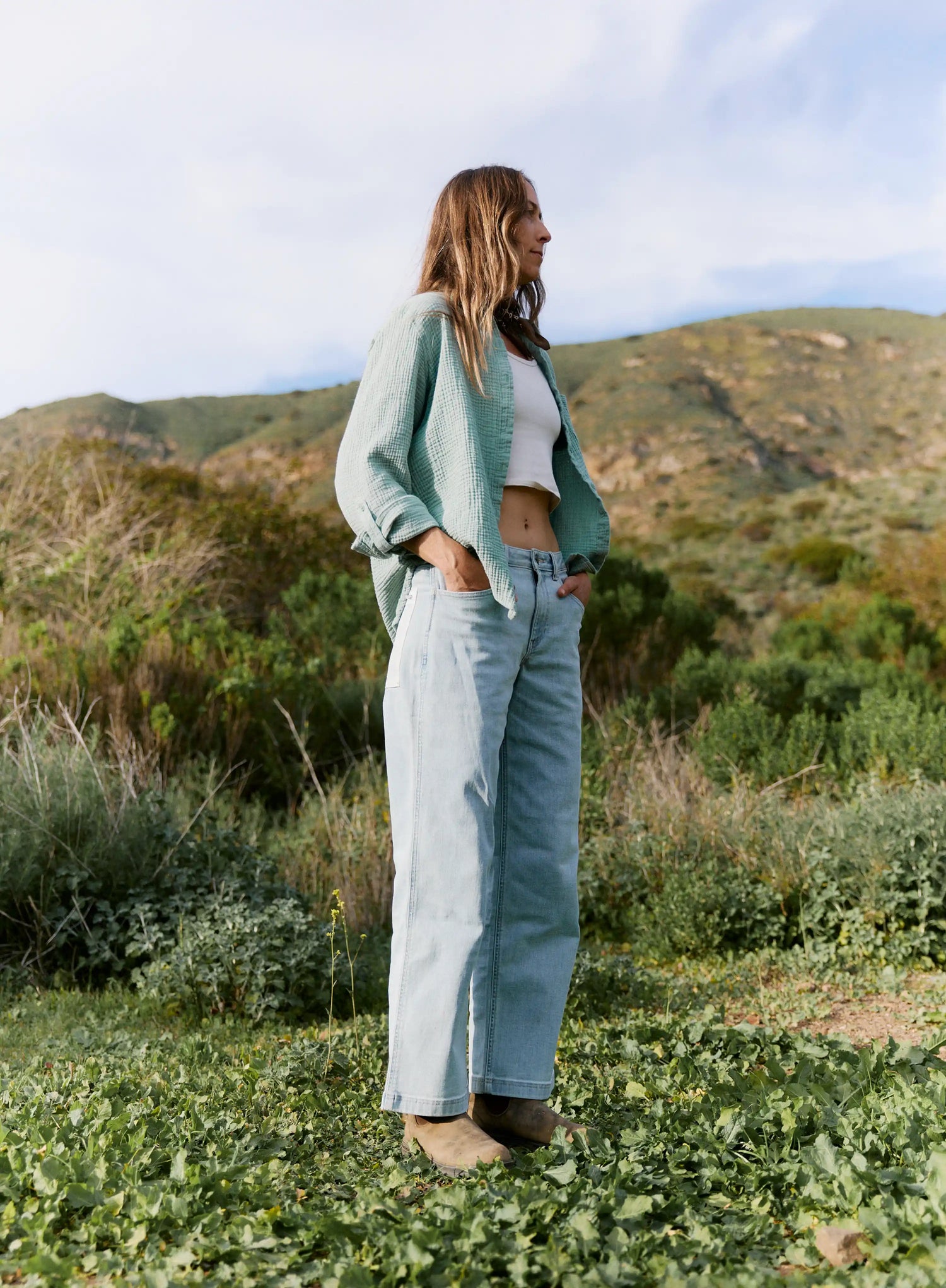 Person wearing a light green jacket and blue jeans standing in a natural landscape with mountains in the background.