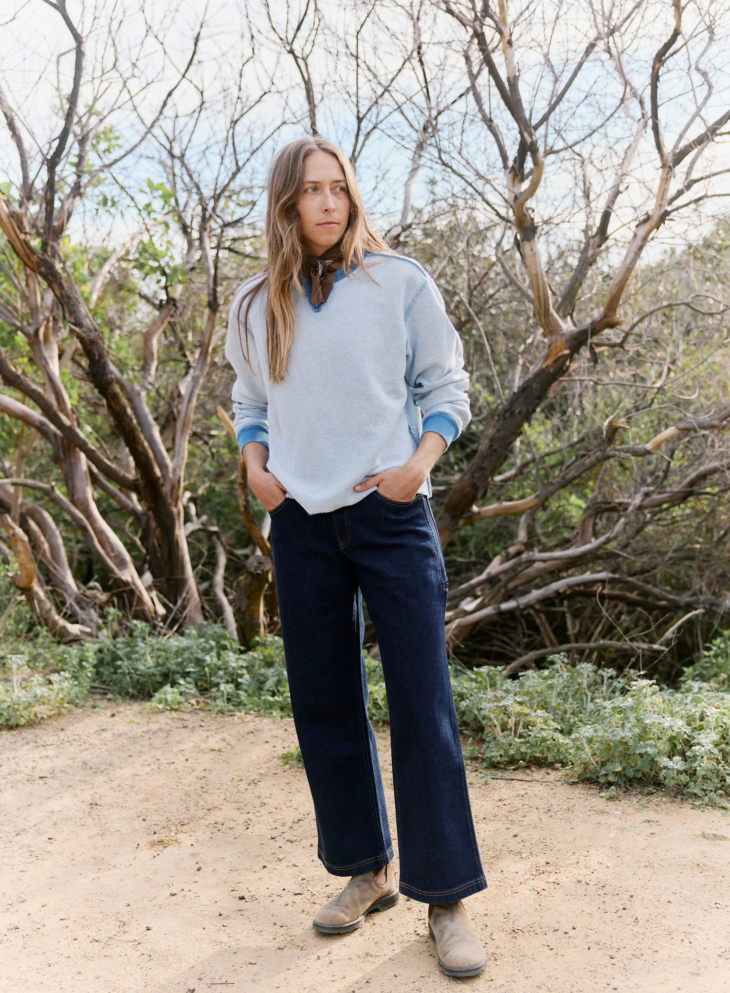 Person wearing a light blue sweater and dark blue pants standing in a natural setting with trees and shrubs.