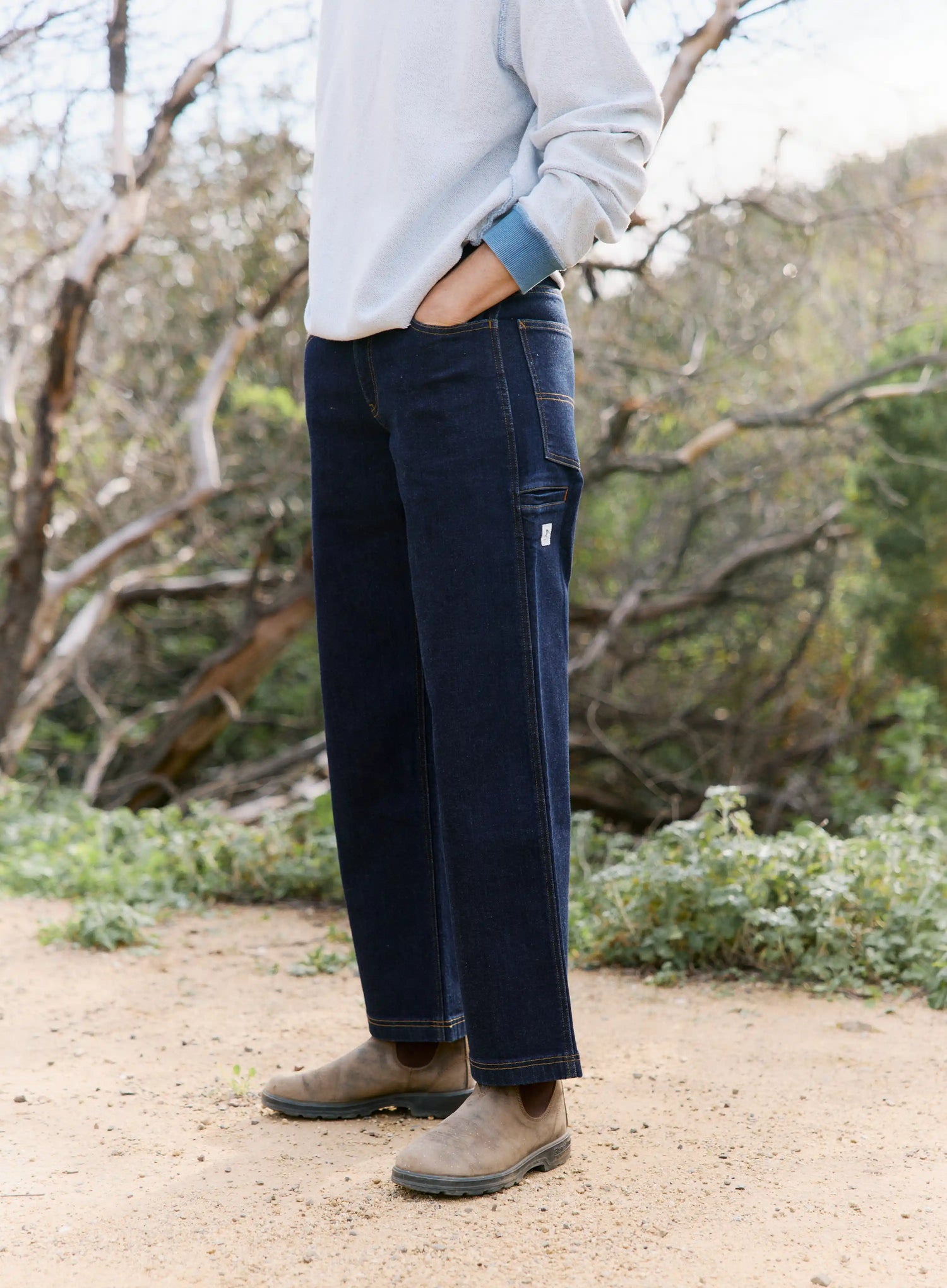 Person wearing dark blue jeans and brown boots standing in a natural setting with trees and shrubs.