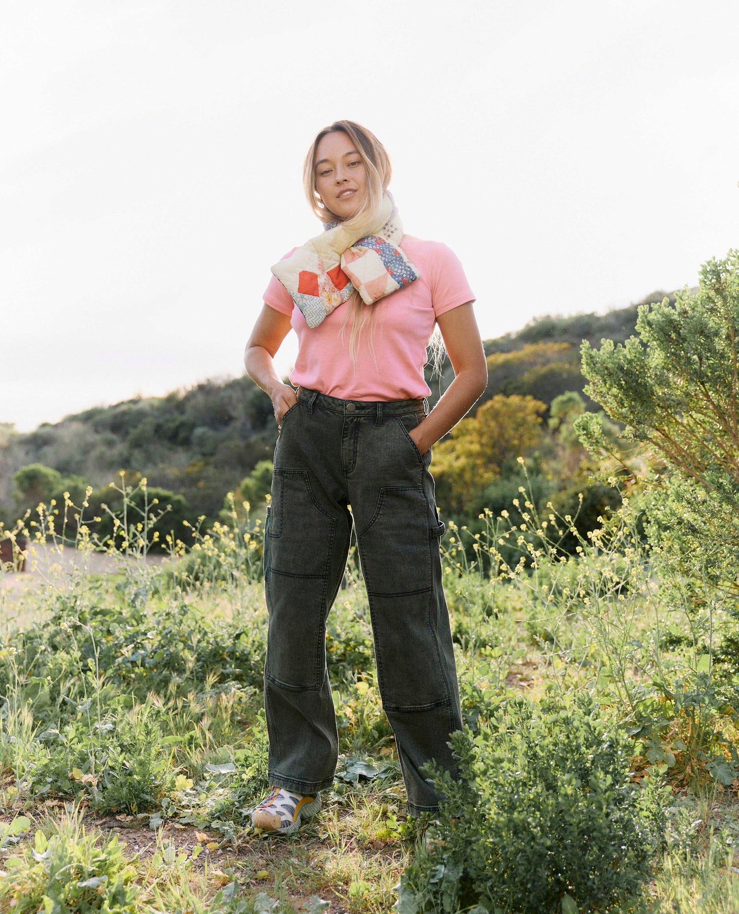 Person wearing a pink shirt and green cargo pants standing in a natural setting with trees and grass.
