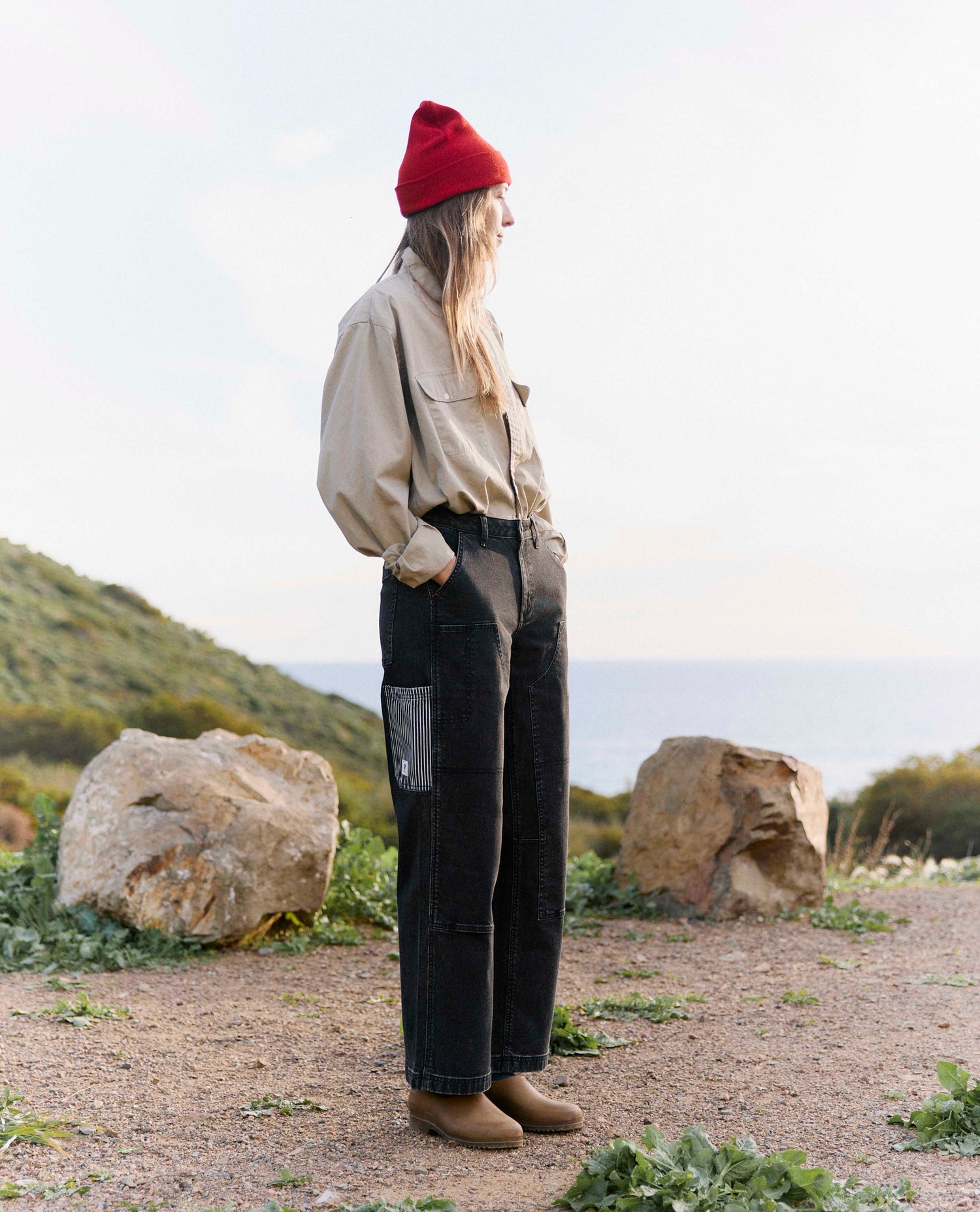 Person wearing a beige jacket, black pants, and a red beanie standing in a natural setting with rocks and grass.