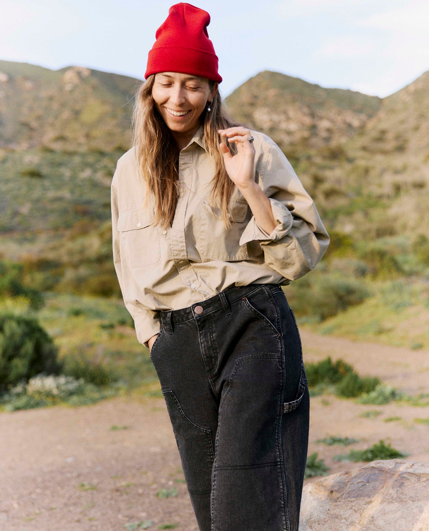 Person wearing a beige shirt, black pants, and a red beanie standing in a natural landscape.