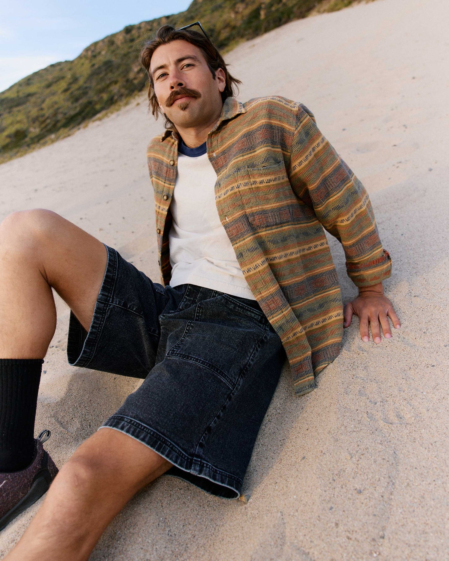 Man sitting on sand wearing a plaid shirt and denim shorts with a scenic background