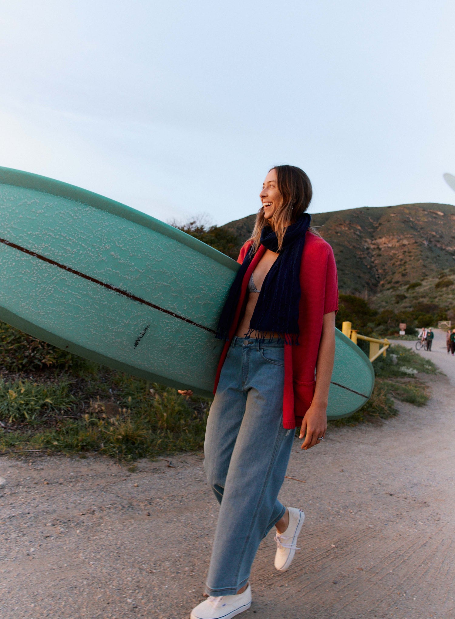 Woman walking with a teal kayak on her shoulder in a scenic outdoor setting.
