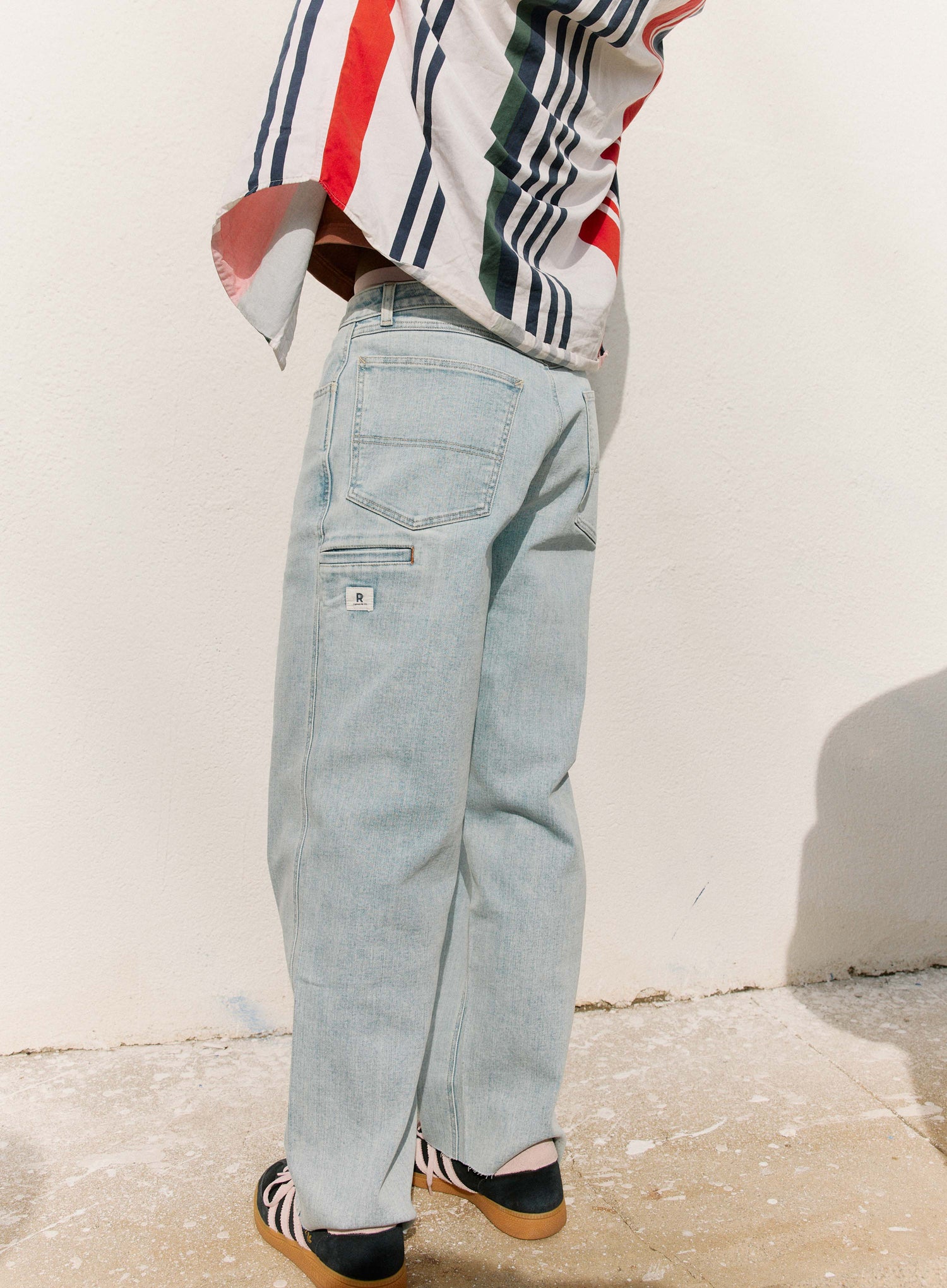 Person wearing a striped shirt and light blue jeans against a white wall.