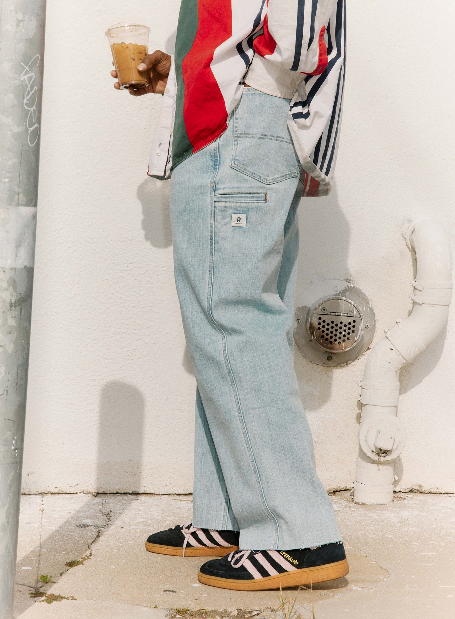 Person wearing light blue jeans and a colorful jacket, standing against a white wall.