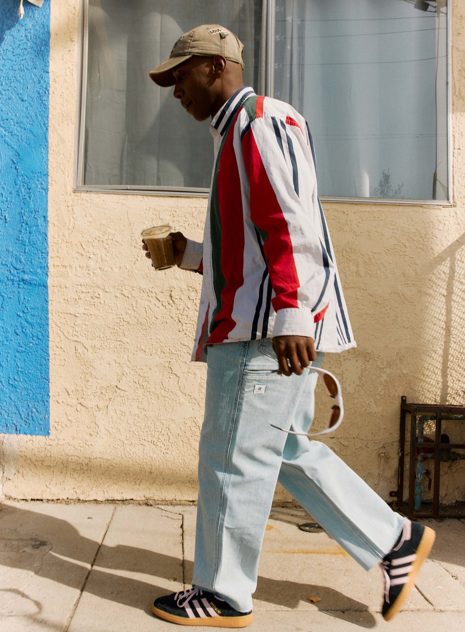 Man walking outdoors holding a coffee cup, wearing a colorful striped shirt and light blue jeans.